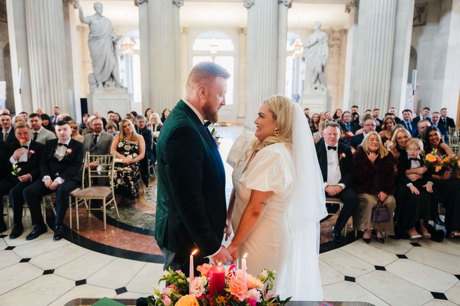 Wedding couple exchanges vows in historic white columned venue surrounded by seated guests. Wedding photographer dublin