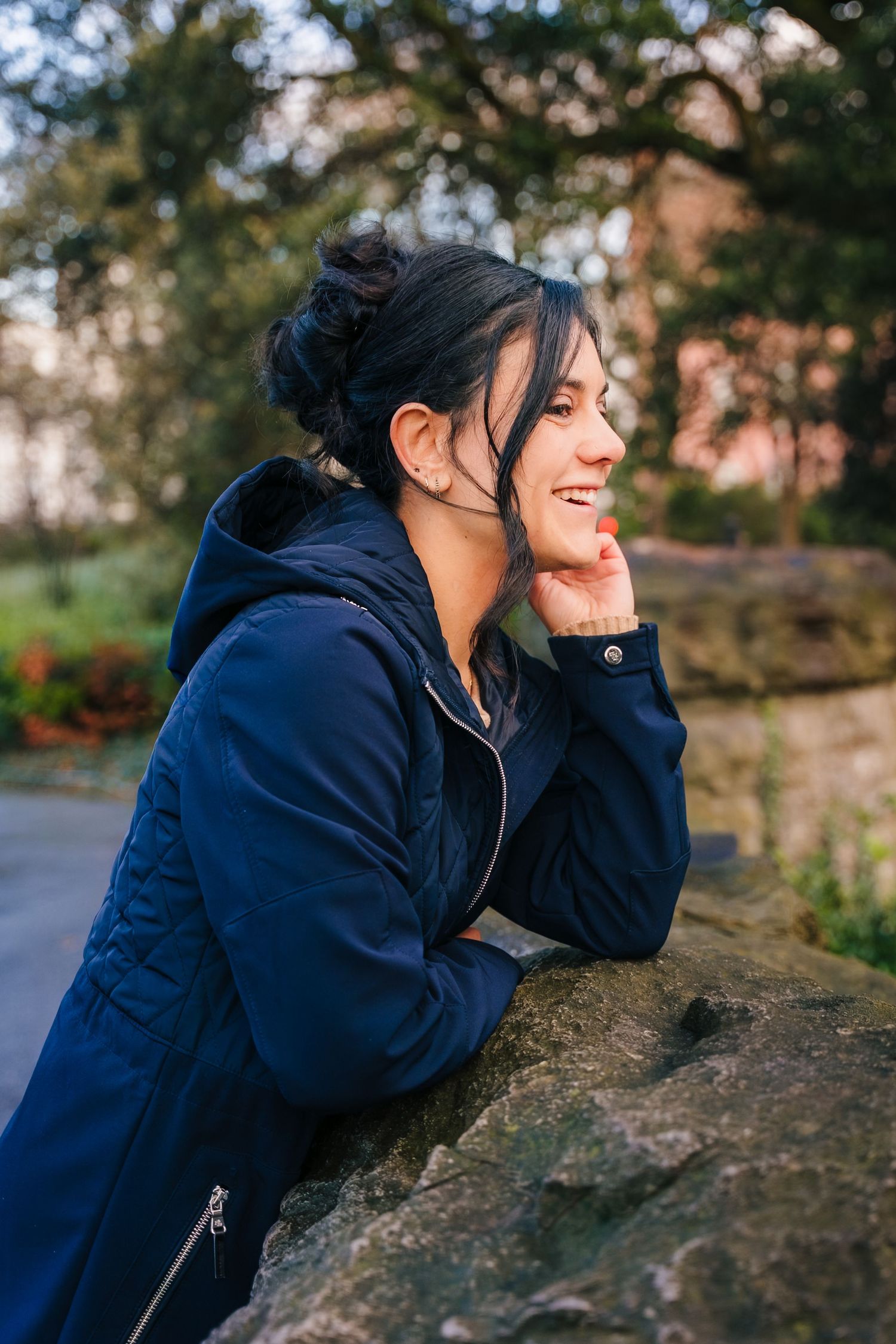 A person in a navy blue hoodie leans on a stone wall while smiling with eyes closed in an outdoor setting. professional headshots dublin