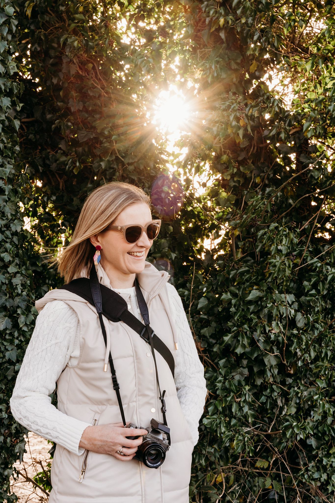 A photographer wearing sunglasses and a white puffy vest holds a camera while standing in sunlit greenery. PHOTOGRAPHER Dublin ireland