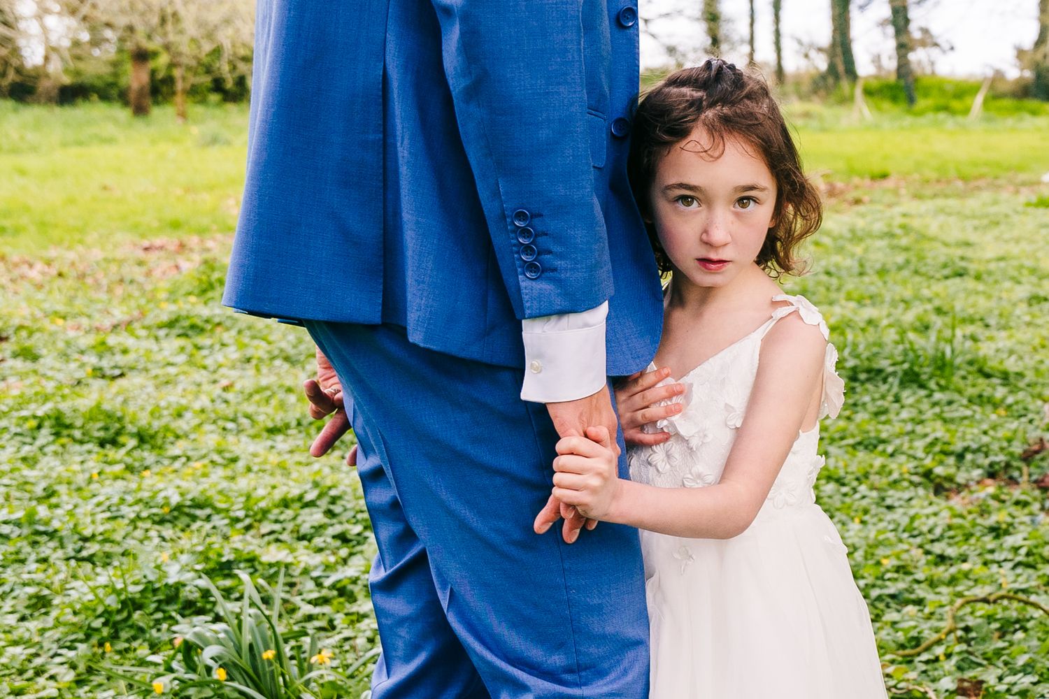 A person in a white dress holds the hand of someone wearing a blue suit in a grassy outdoor setting. family PHOTOGRAPHER Dublin