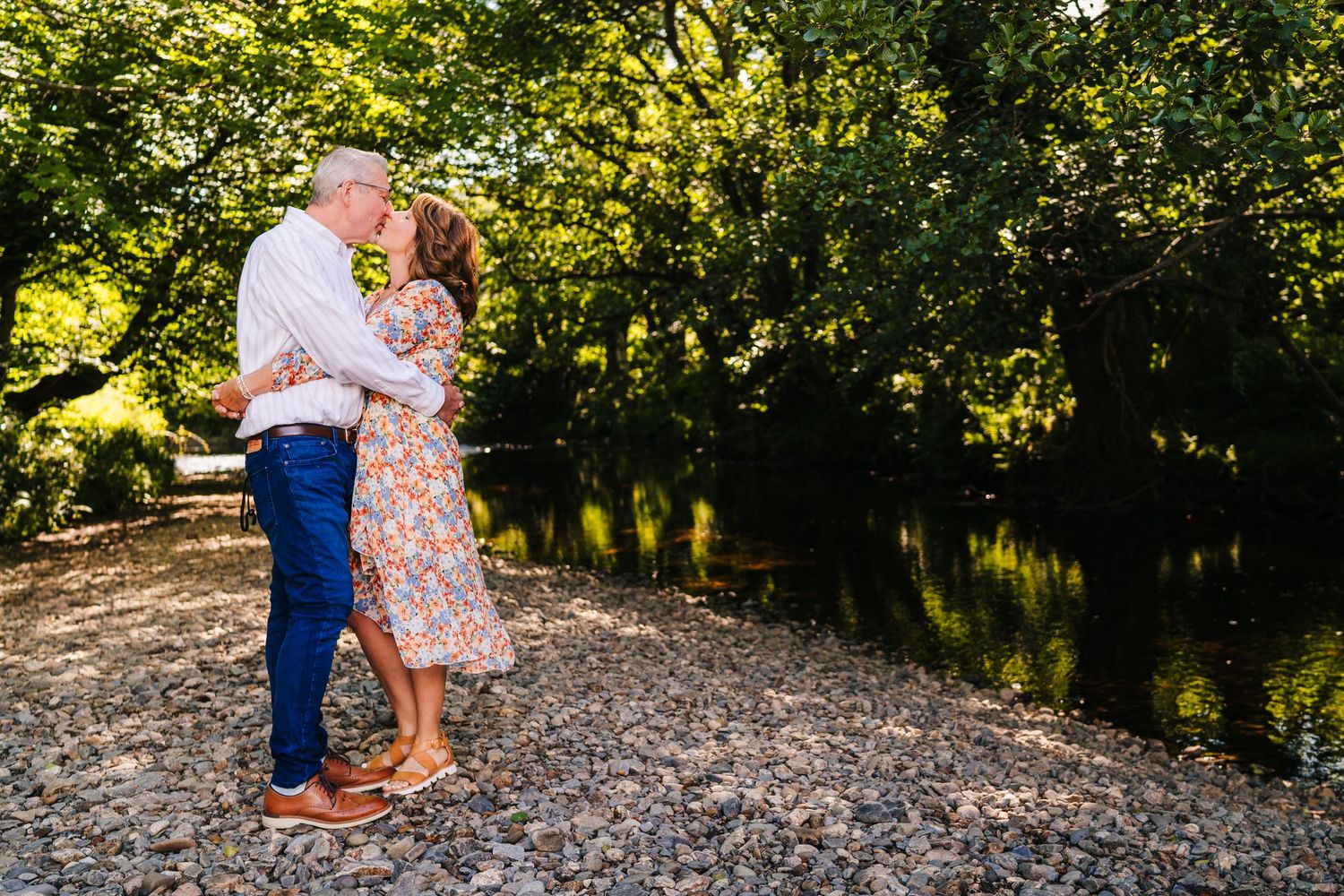 A romantic couple embraces by a tranquil riverbank on a sunny day amid lush green trees and foliage. PHOTOGRAPHER Dublin ireland
