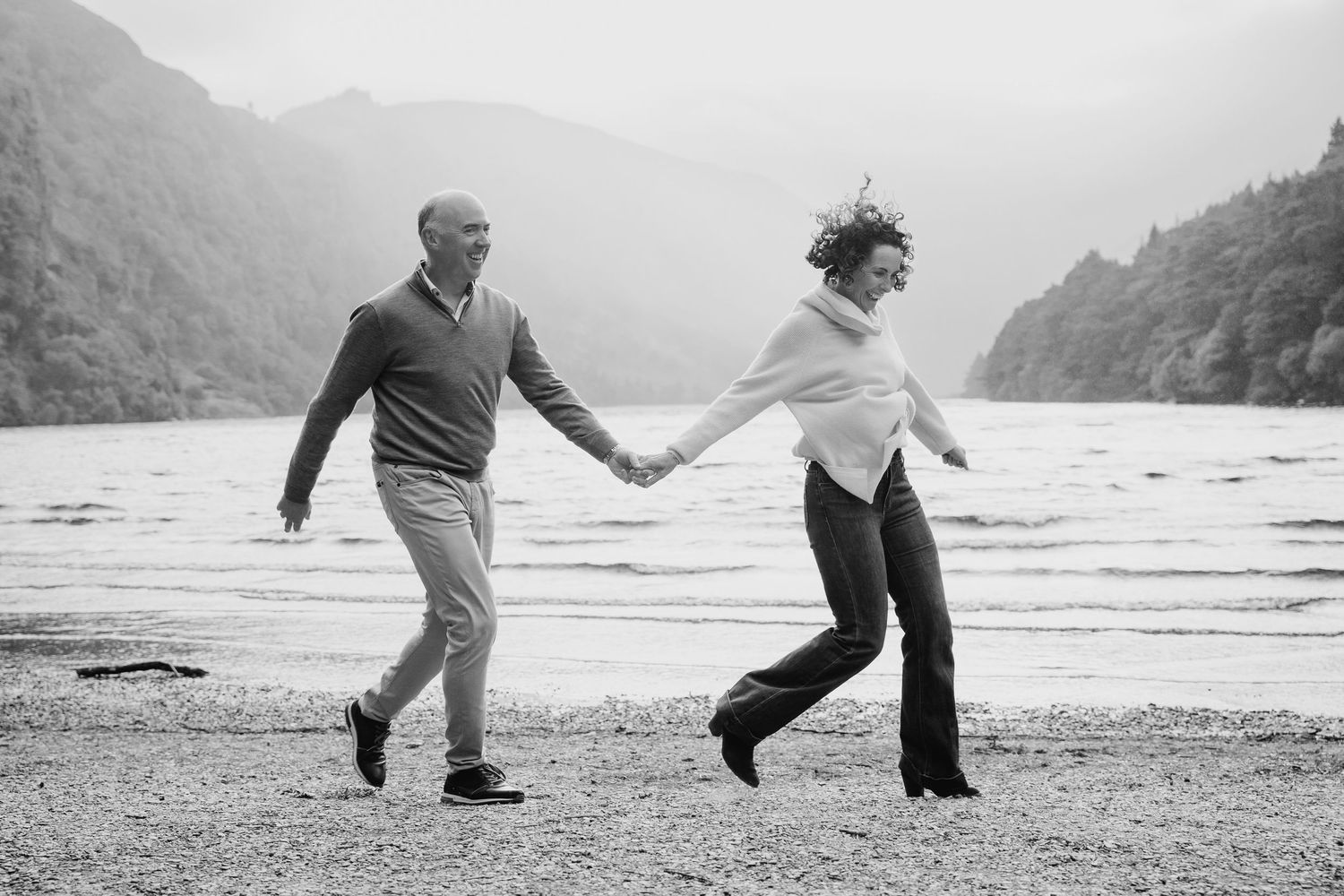 Black and white photo of a couple dancing and holding hands on a beach with mountains in the distance. PHOTOGRAPHER Dublin ireland