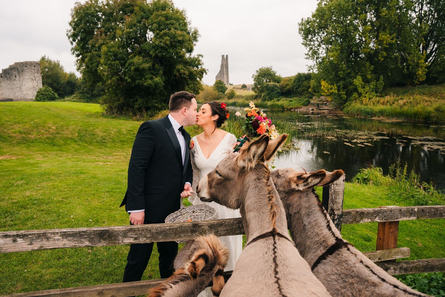 Couple poses with a donkey in a rustic outdoor setting with green fields and trees in the background. Wedding PHOTOGRAPHER Dublin