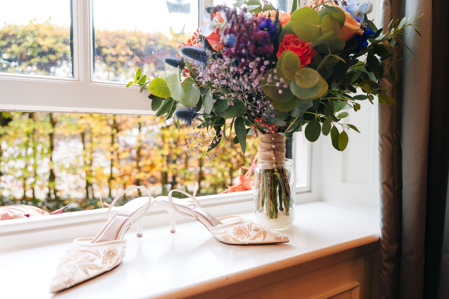 A colorful floral arrangement in a glass vase sits on a windowsill overlooking autumn trees outside.