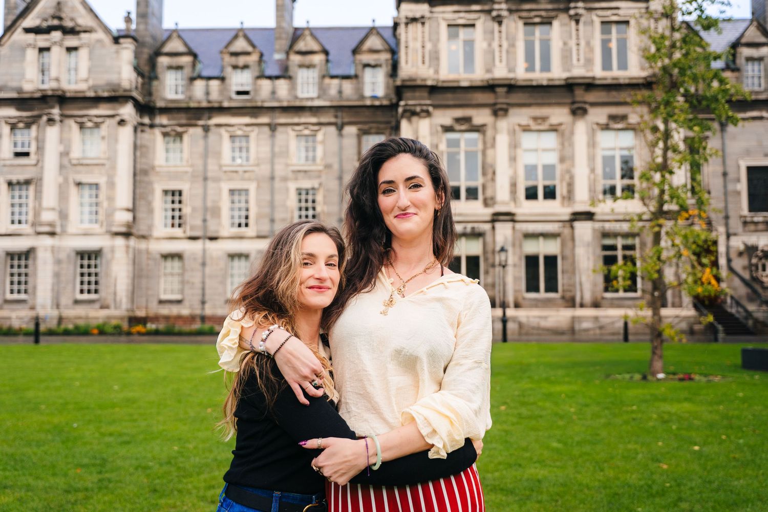 A couple poses together on a grassy lawn in front of a grand historic stone building. professional headshots dublin photographer