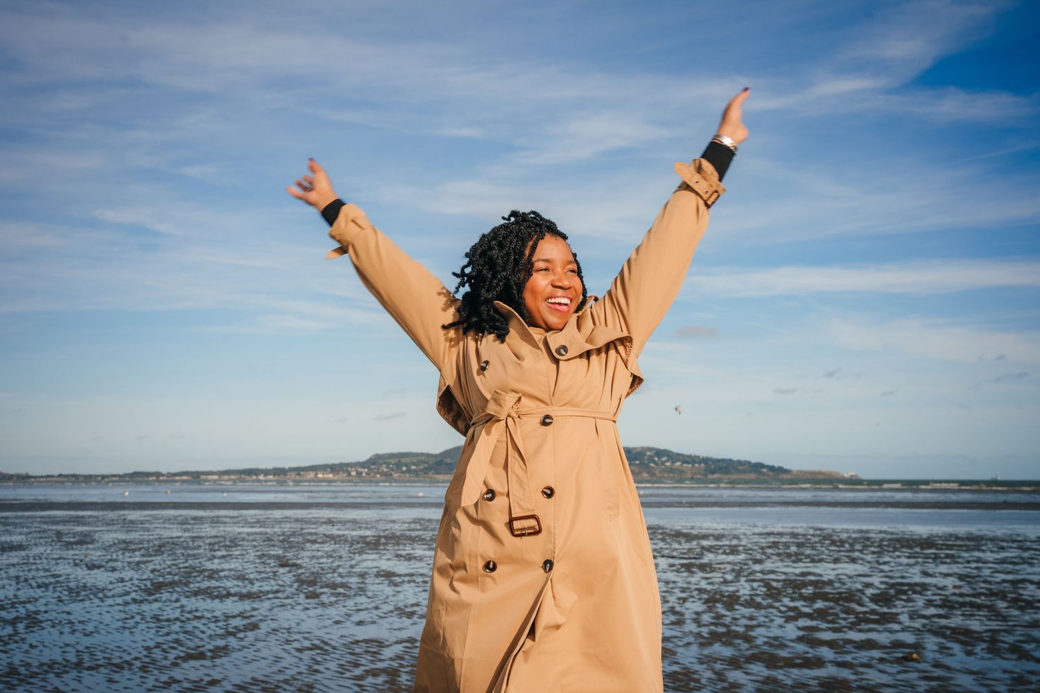 Person in tan coat expressing joy with raised arms against blue sky and coastal landscape. Dublin professional headshot photographer
