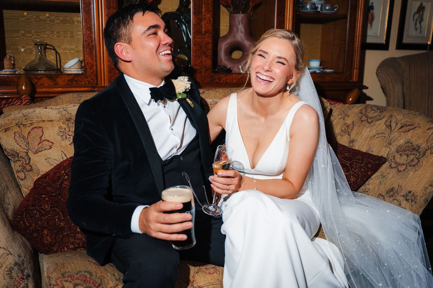 Wedding couple shares intimate moments and laughter while enjoying drinks on an elegant floral couch. Ballymagarvey Wedding photographer