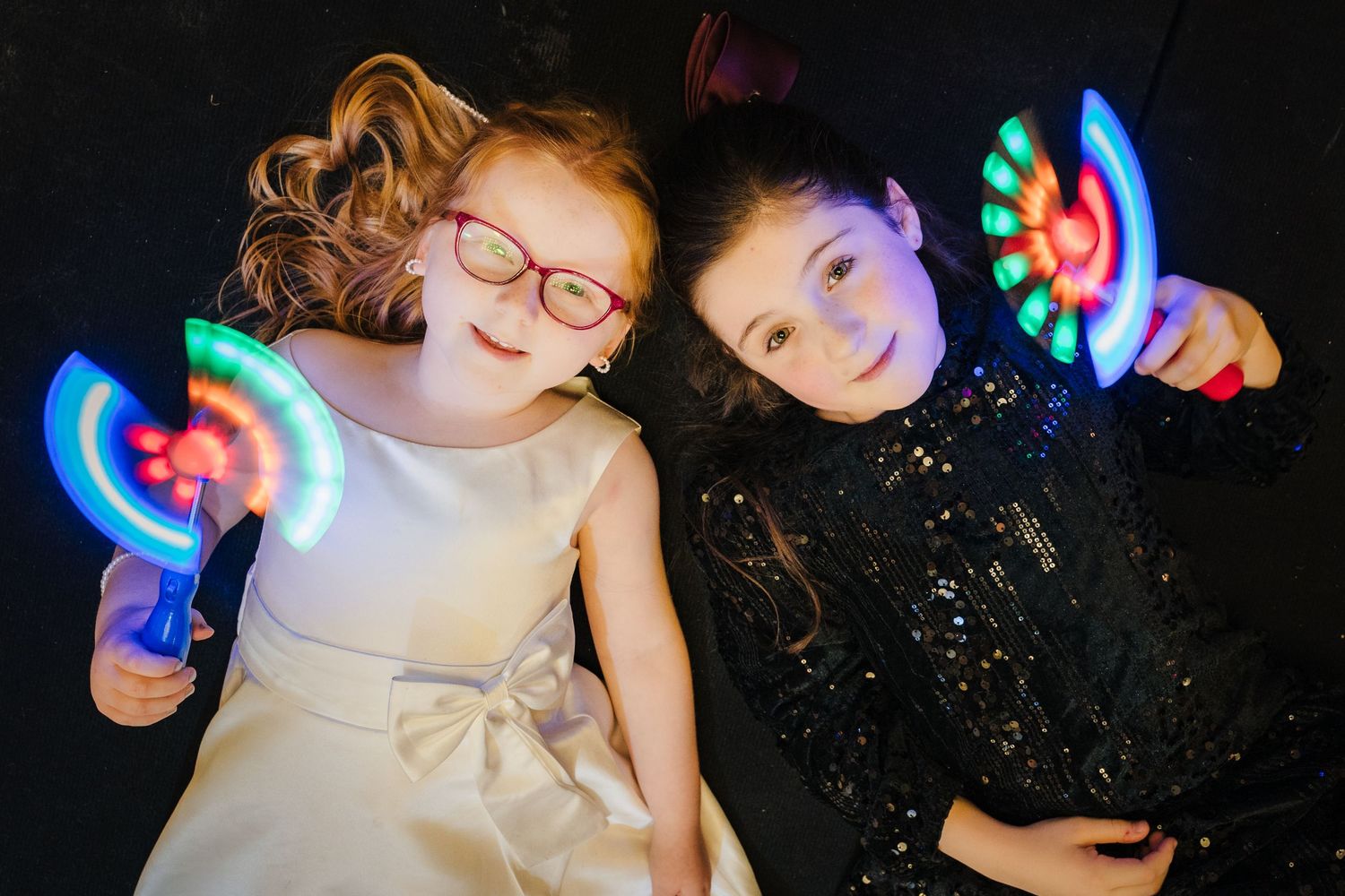 Two children lie on floor holding colorful LED light-up toys creating a playful nighttime scene.  Wedding PHOTOGRAPHER Trim Castle