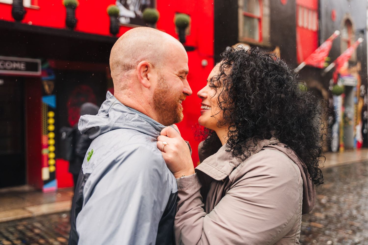 People sharing an umbrella while walking past red market stalls on a wet cobblestone street. Proposal Dublin Photographer