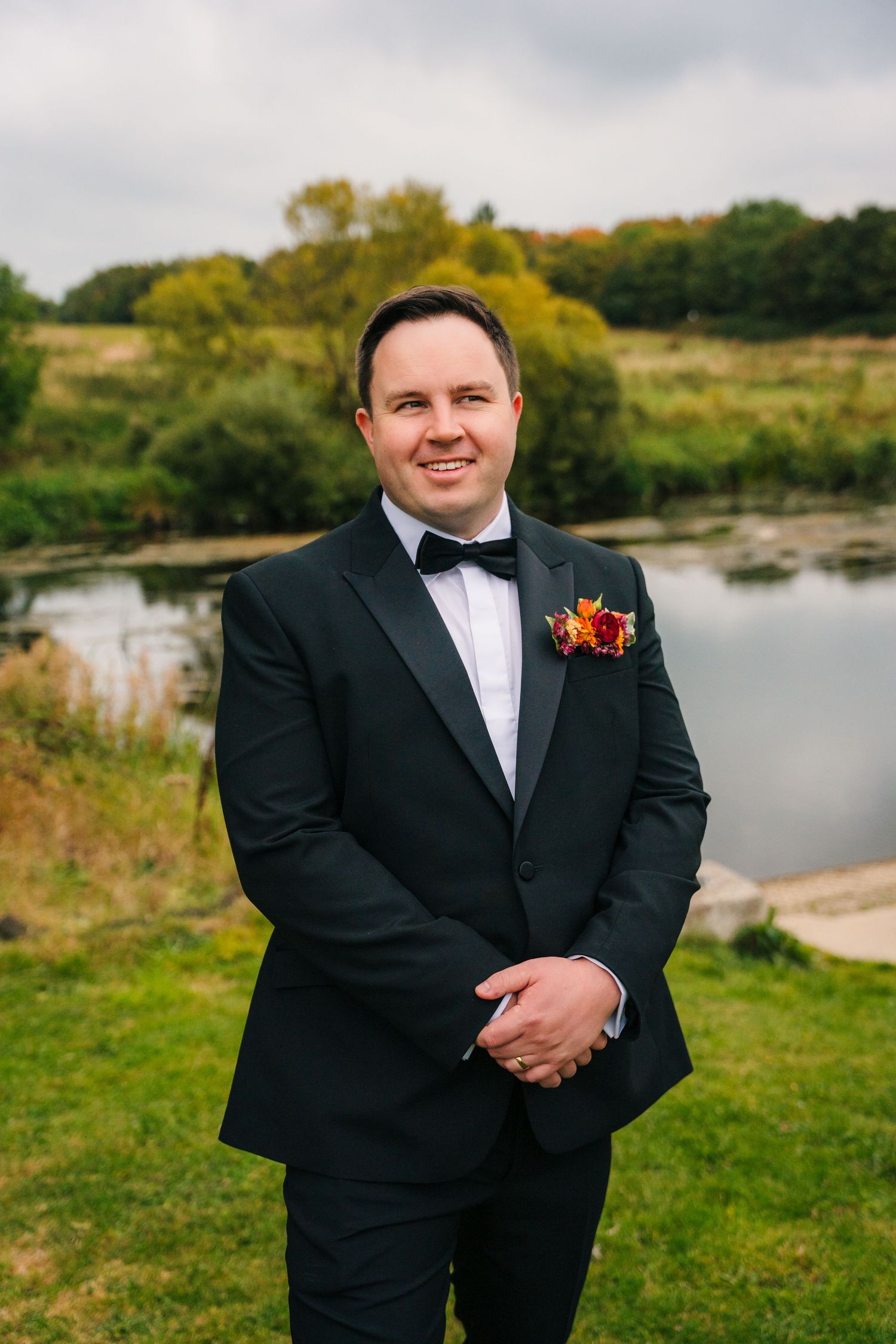 Groom in classic black tuxedo poses for portraits near a scenic lake and grassy landscape.Wedding Photography Trim Castle Ireland