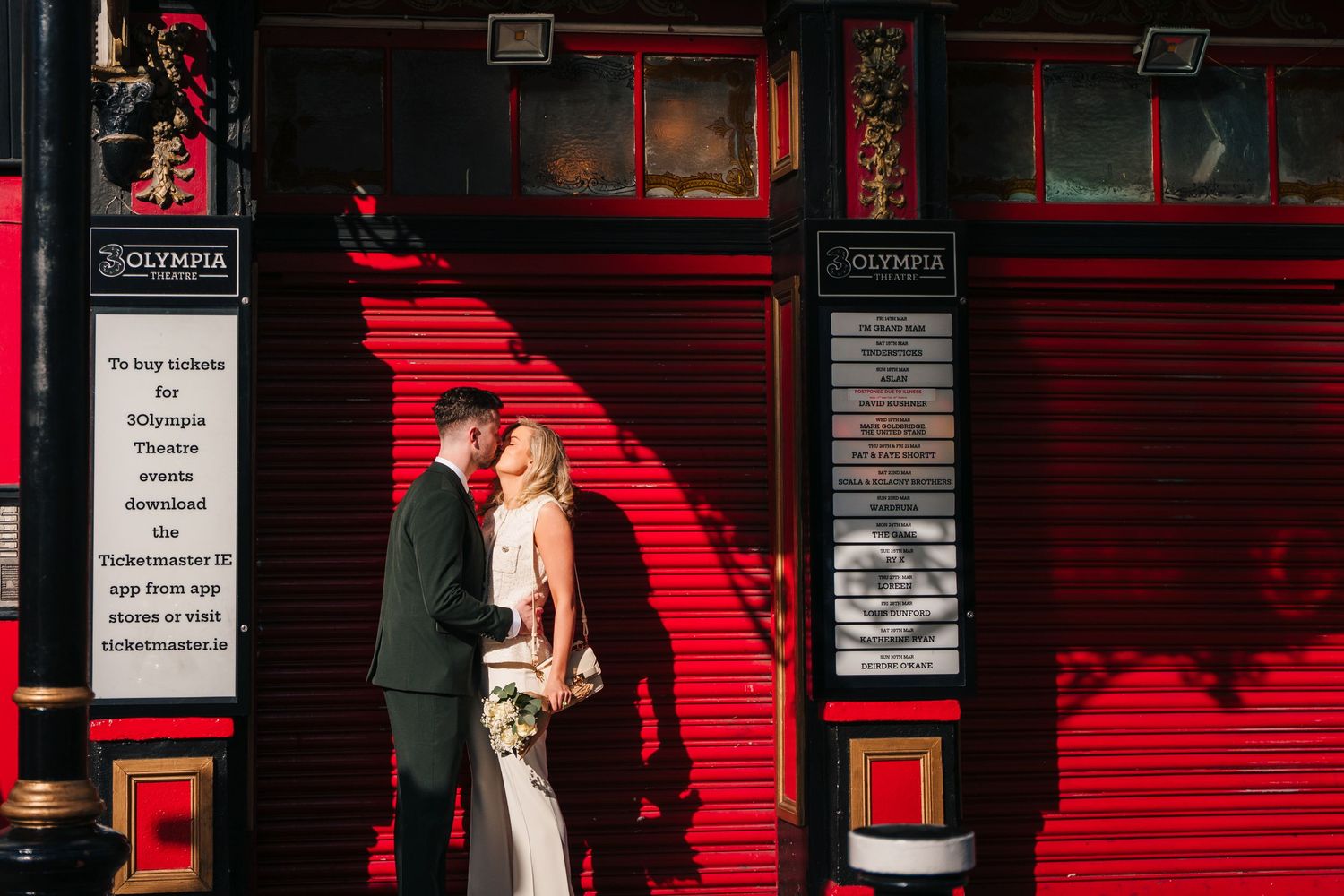 Dramatic sunlit photo of couple against bright red storefront with striking shadows. City Hall Dublin wedding photogrAPher