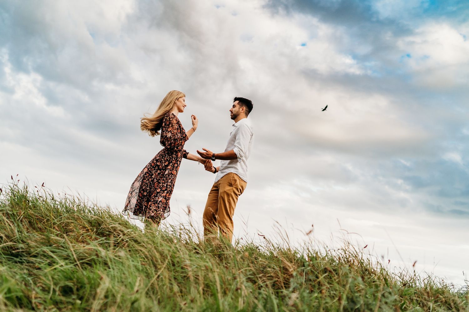 Couple holds hands while walking through grassy field at sunset under dramatic cloudy sky. PHOTOGRAPHER Dublin ireland