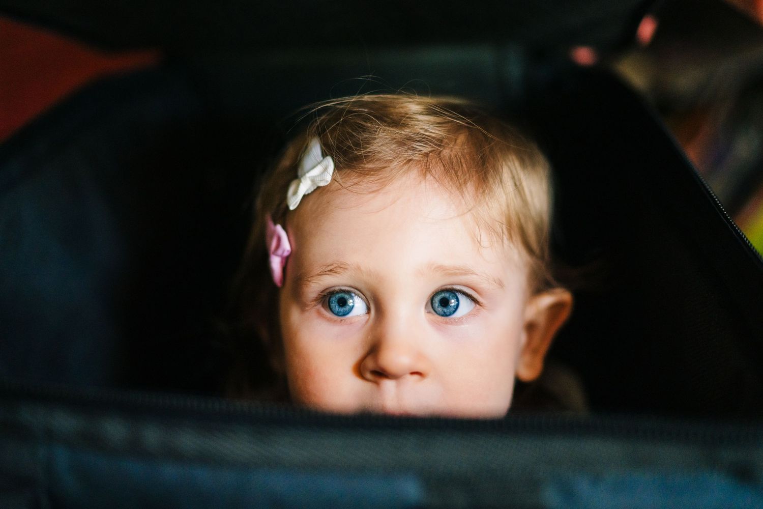 A small child with bright blue eyes peeks out from behind a seat, wearing a white hair clip. family PHOTOGRAPHER Dublin