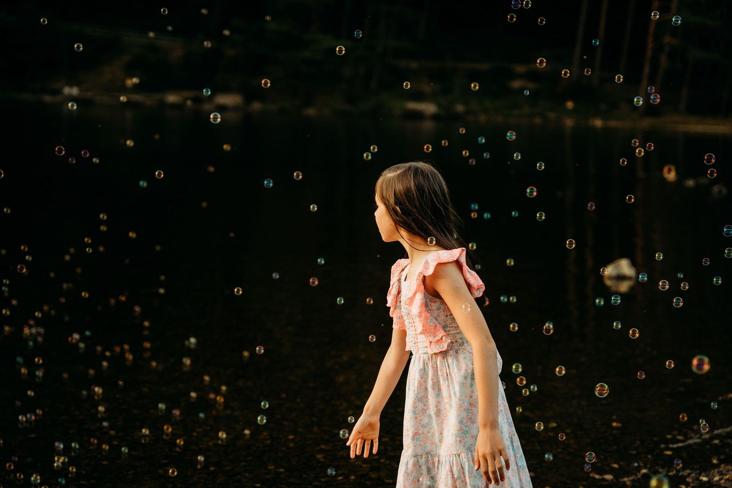 A child in a white summer dress plays with bubbles in a dark backyard during golden hour. family PHOTOGRAPHER Dublin
