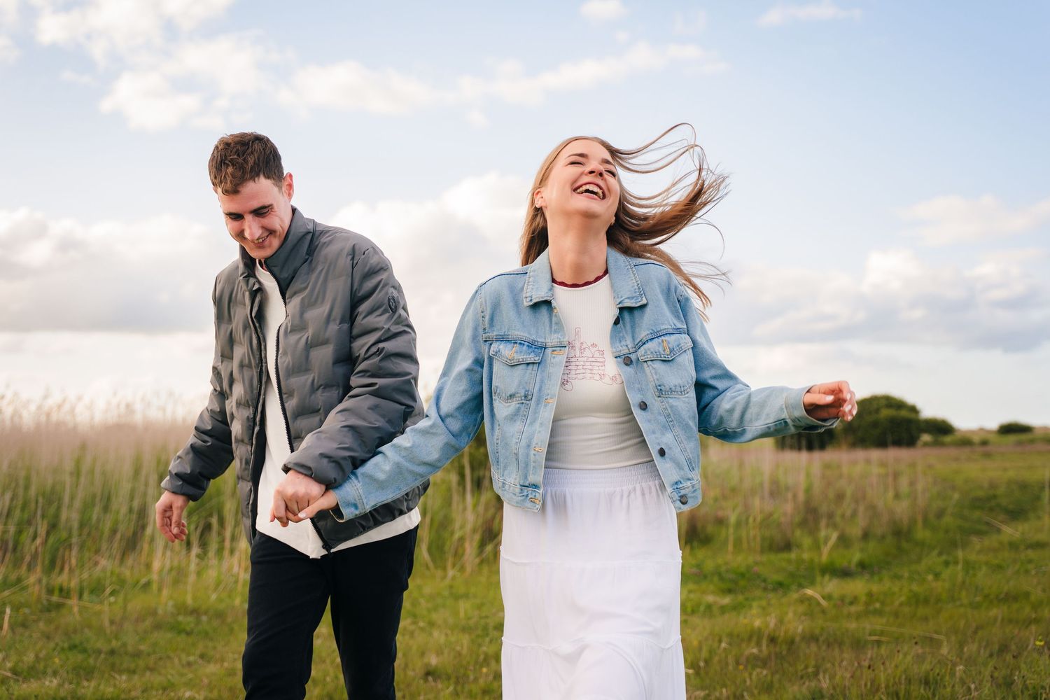 A couple laughs and embraces while standing in a sunny field on a cloudy day. PHOTOGRAPHER Dublin ireland