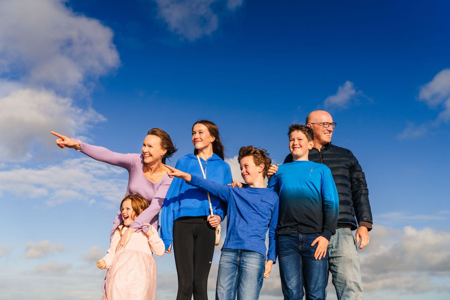 Group wearing blue stands together pointing at sky against fluffy white clouds on bright sunny day. Family PHOTOGRAPHER Dublin