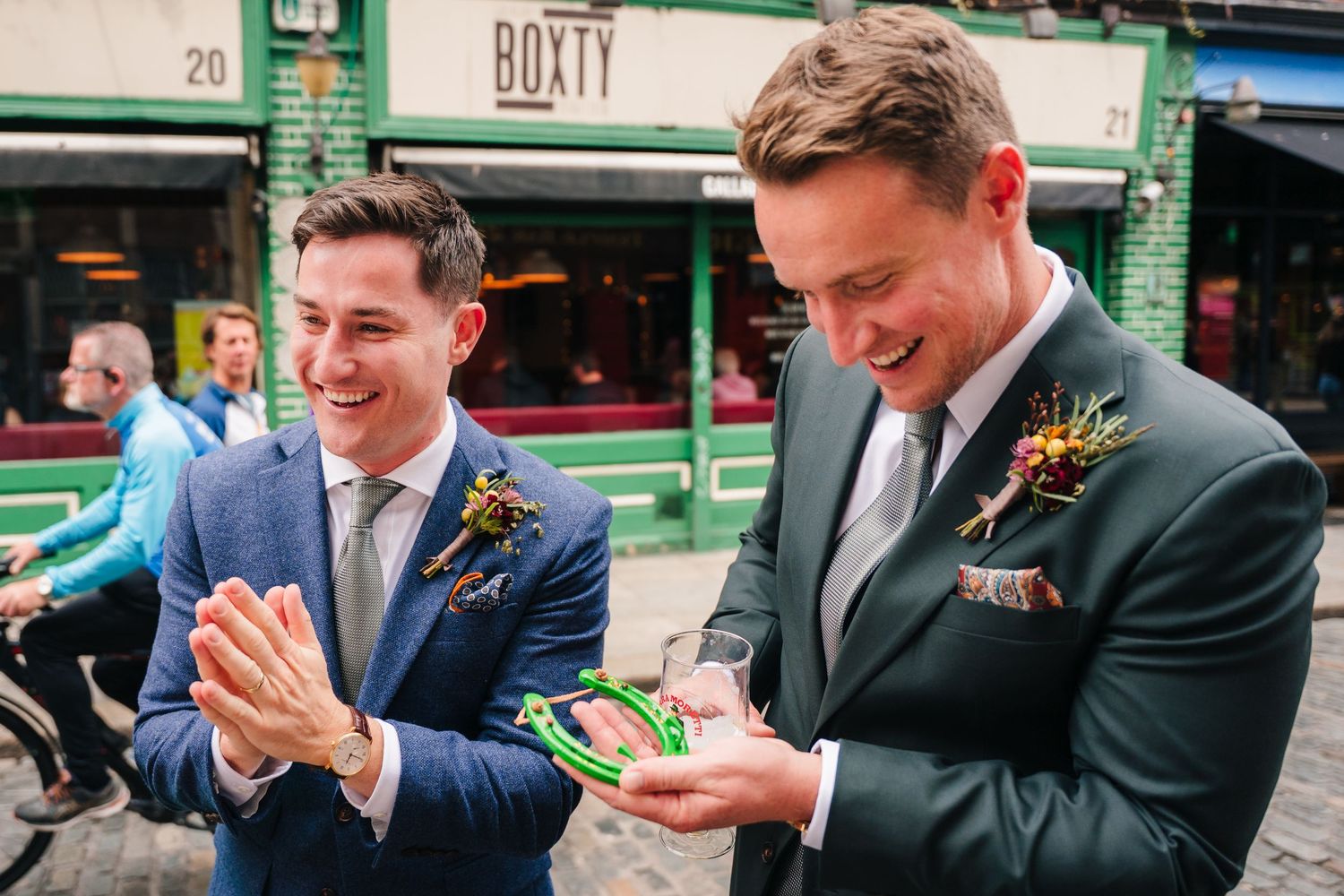 Two wedding guests in blue and black suits share a joyful moment together on the street while wearing boutonnieres. Same sex wedding PHOTOGR