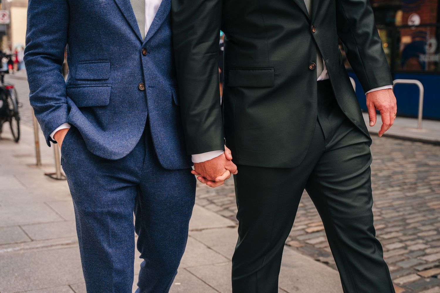 Two people wearing suits walk together on a cobblestone street in a city setting. Same sex wedding PHOTOGRAPHER Dublin