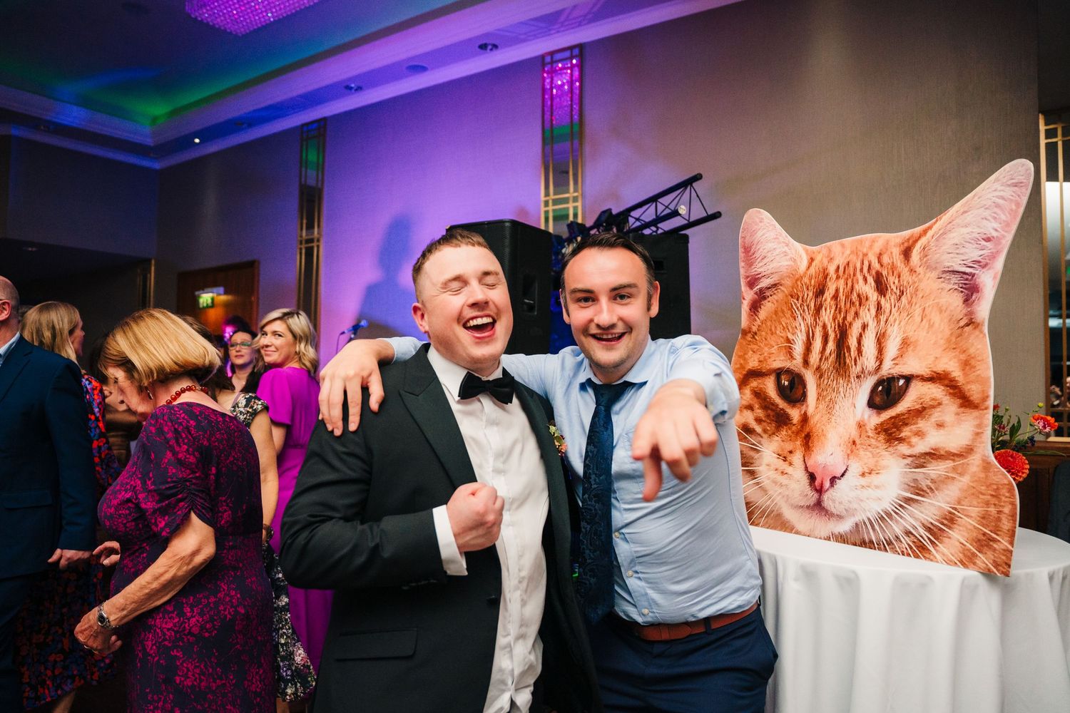 Wedding reception guests pose with large cat cutout prop amid festive purple lighting.Wedding Photography Trim Castle Ireland