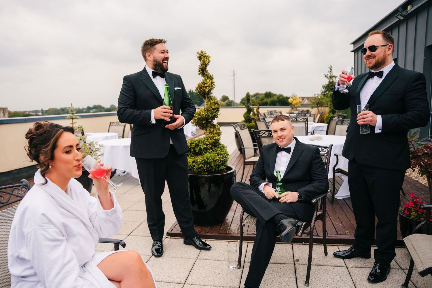 A group in formal attire enjoying drinks and conversation on an outdoor rooftop patio with decorative topiary. Wedding Photography Trim Cast