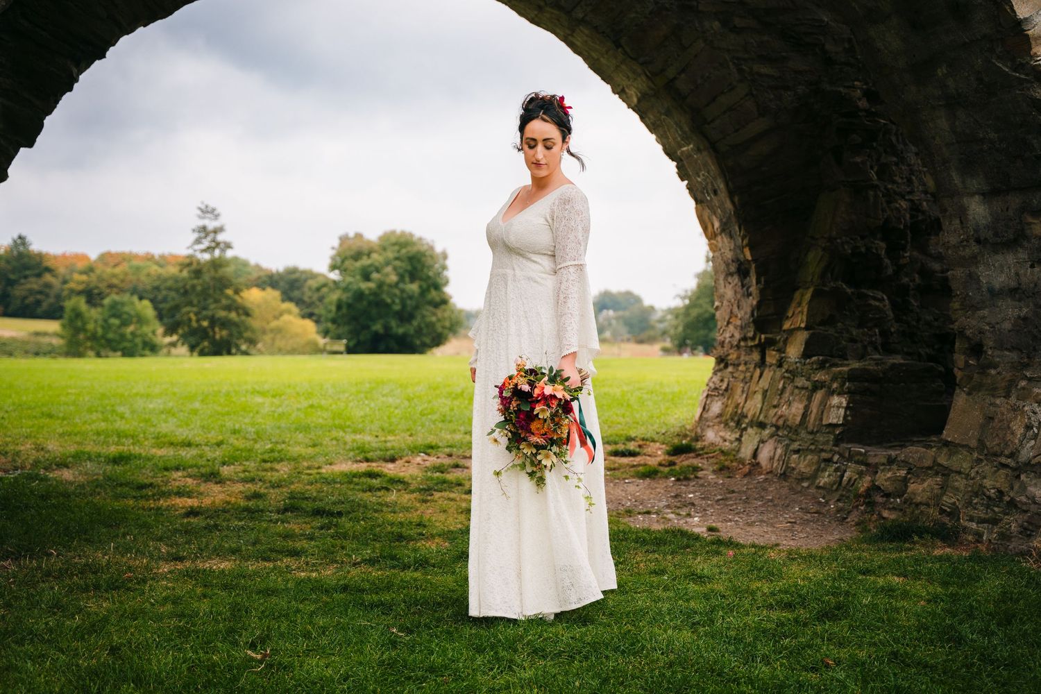 A person in a wedding dress poses under a stone arch in a pastoral countryside setting. Wedding Photography Trim Castle Ireland