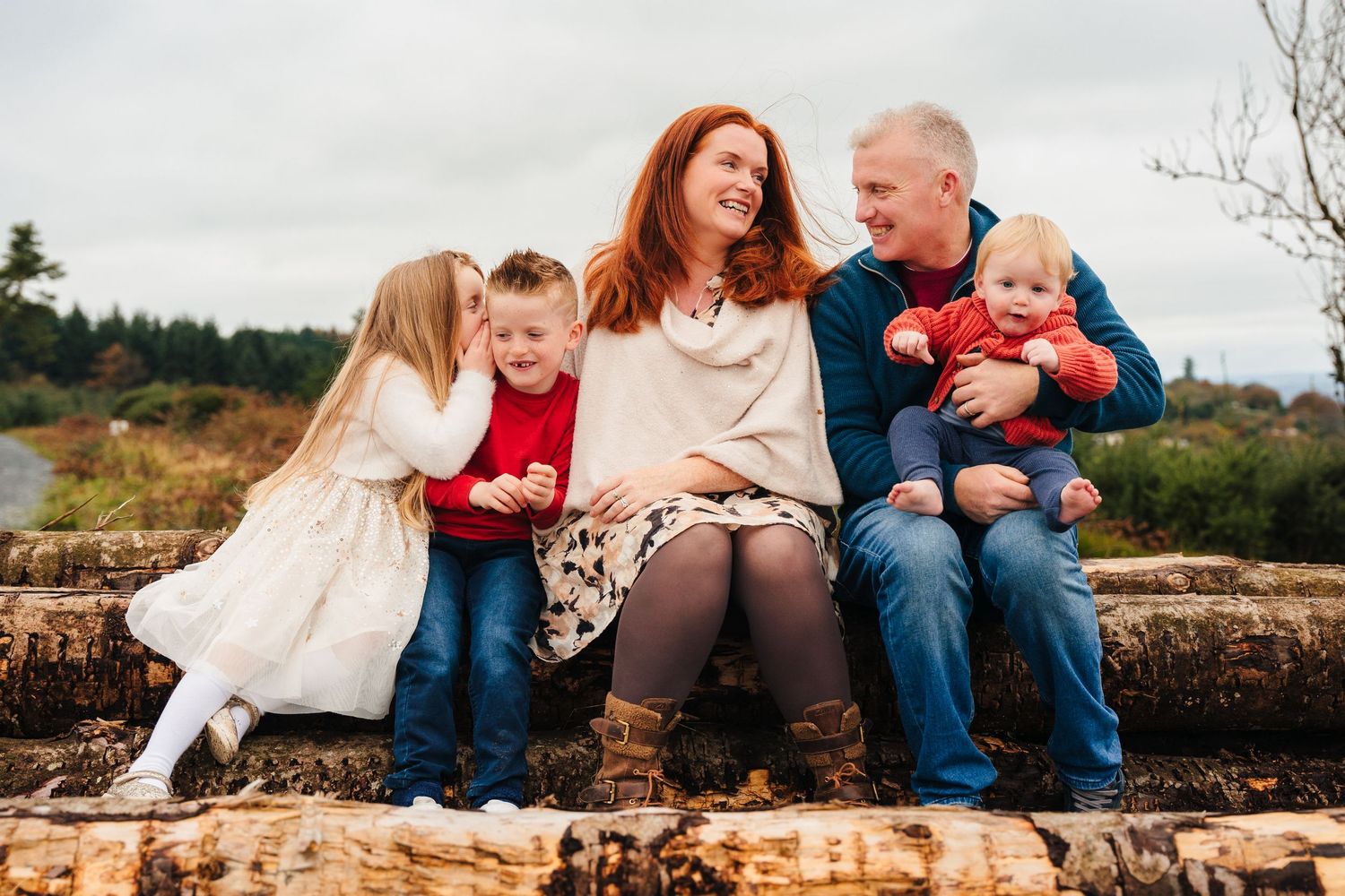 A family of five sitting together and laughing on a large fallen log in a natural outdoor setting. Family Photographer Dublin Ireland