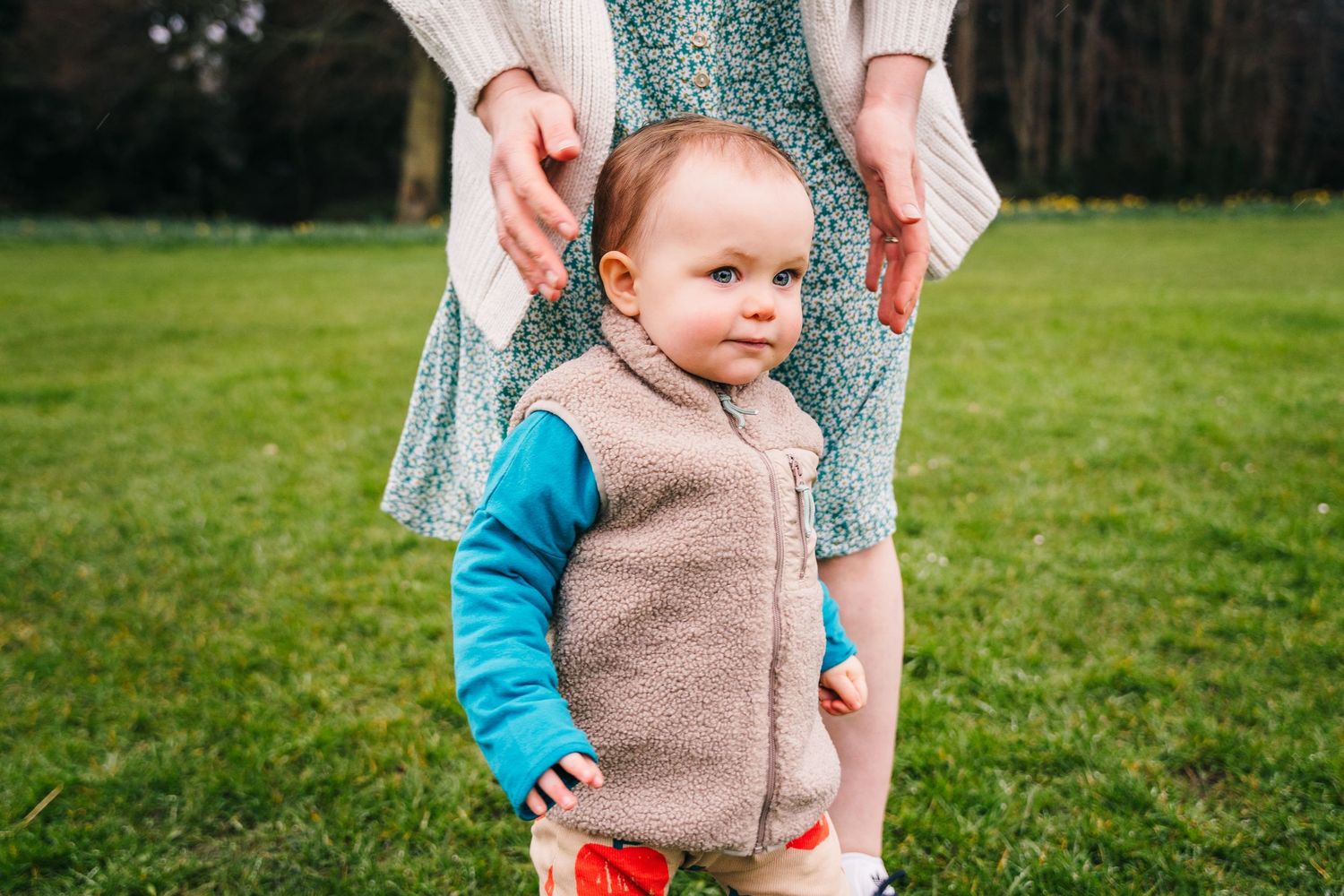 Family photographer Dublin Ireland A small child in a beige vest stands in a grassy field while being steadied by adult hands.