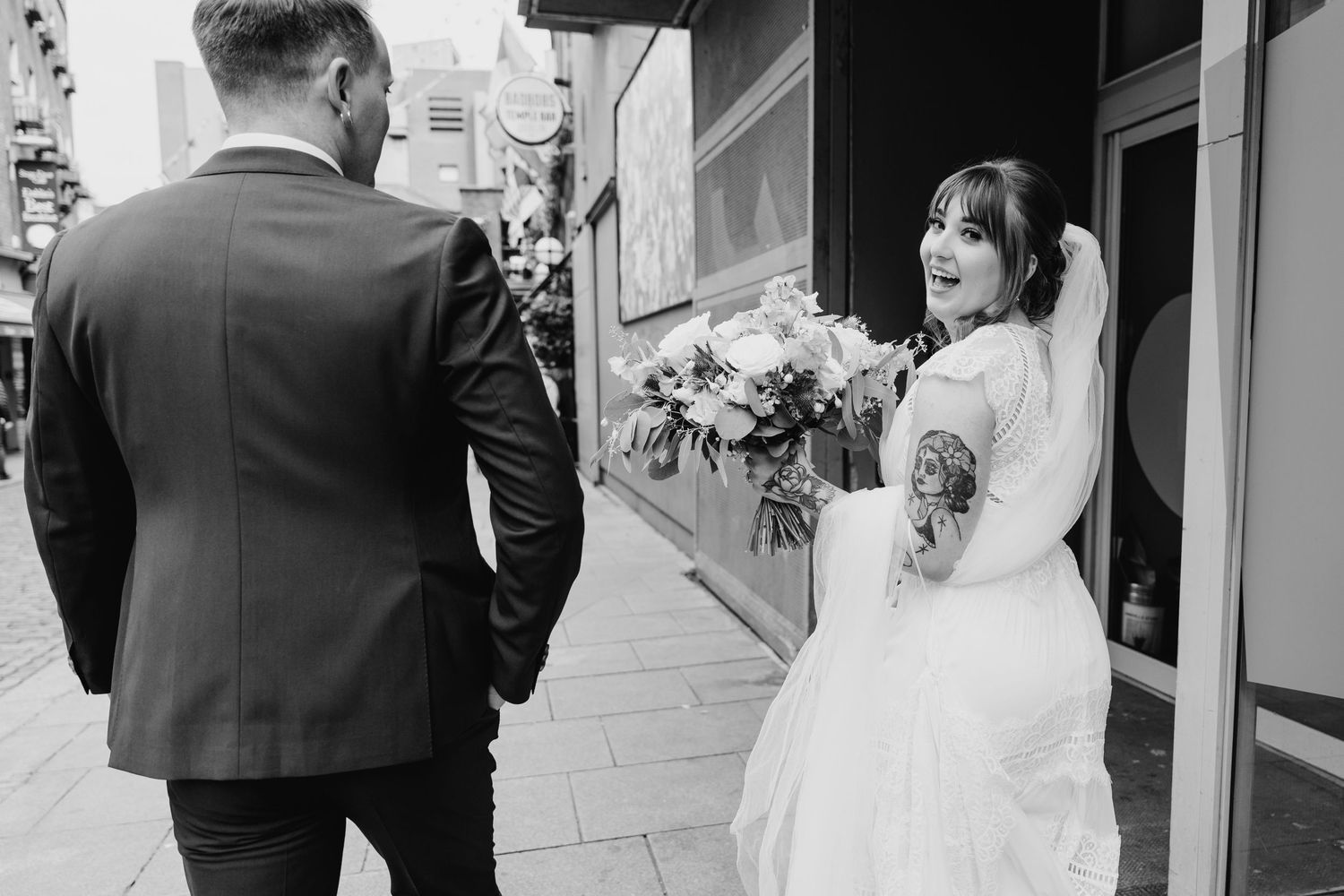 Black and white photo of bride with tattoo reacting joyfully on city sidewalk. PHOTOGRAPHER Dublin Ireland Wedding