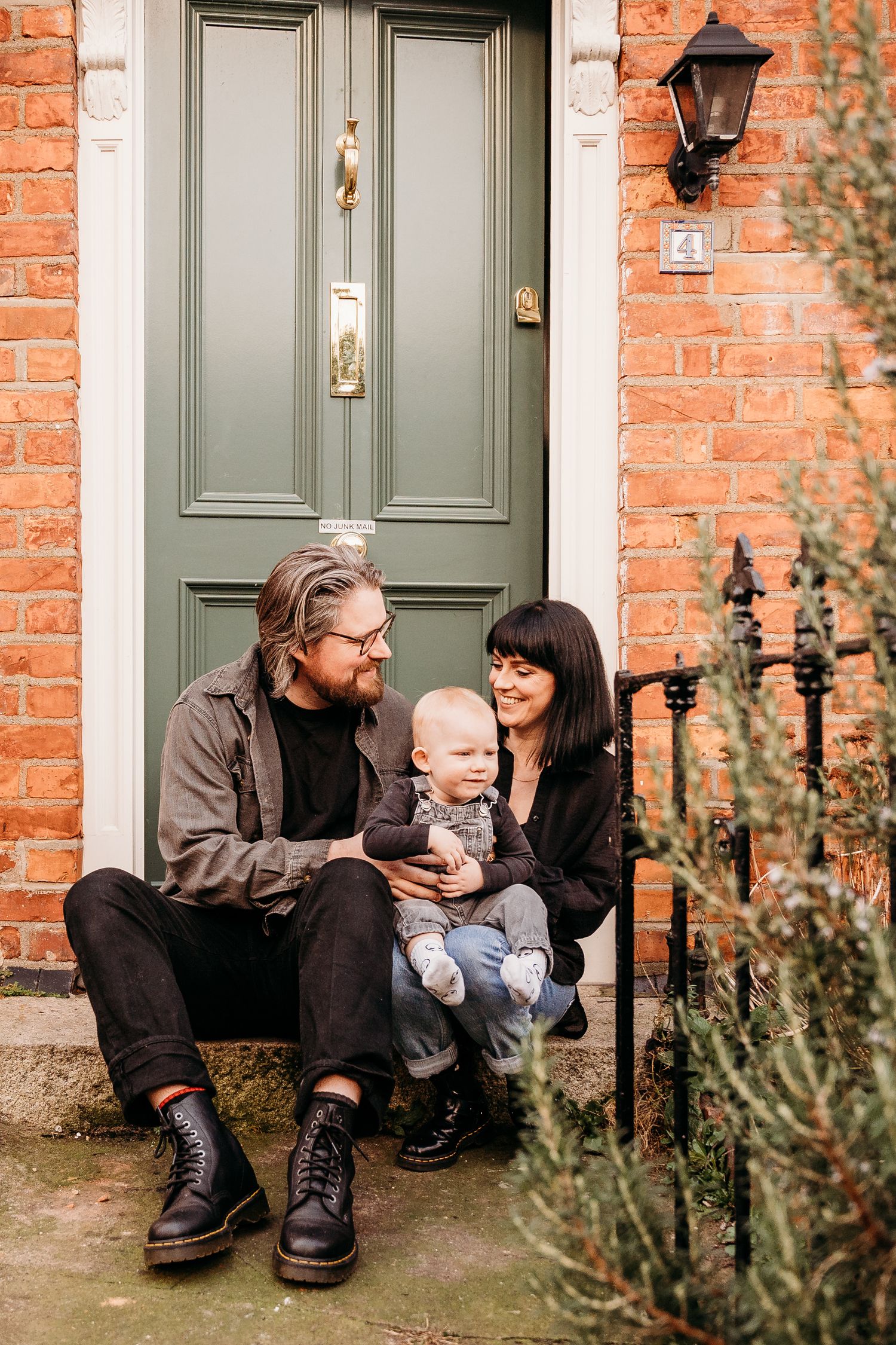 A family sits together on stone steps by a green front door of a brick house with a vintage lantern.  Family PHOTOGRAPHER Dublin