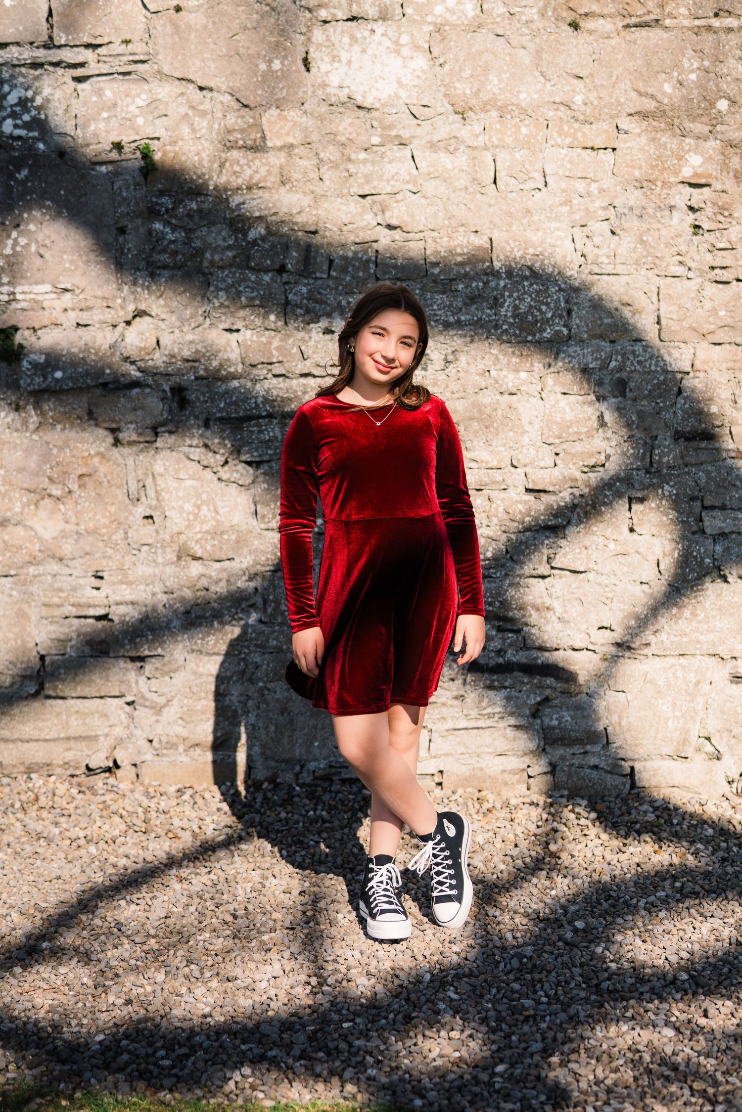 A person wearing a red velvet dress and black sneakers poses against a stone wall with dramatic tree shadows. Family PHOTOGRAPHER Dublin