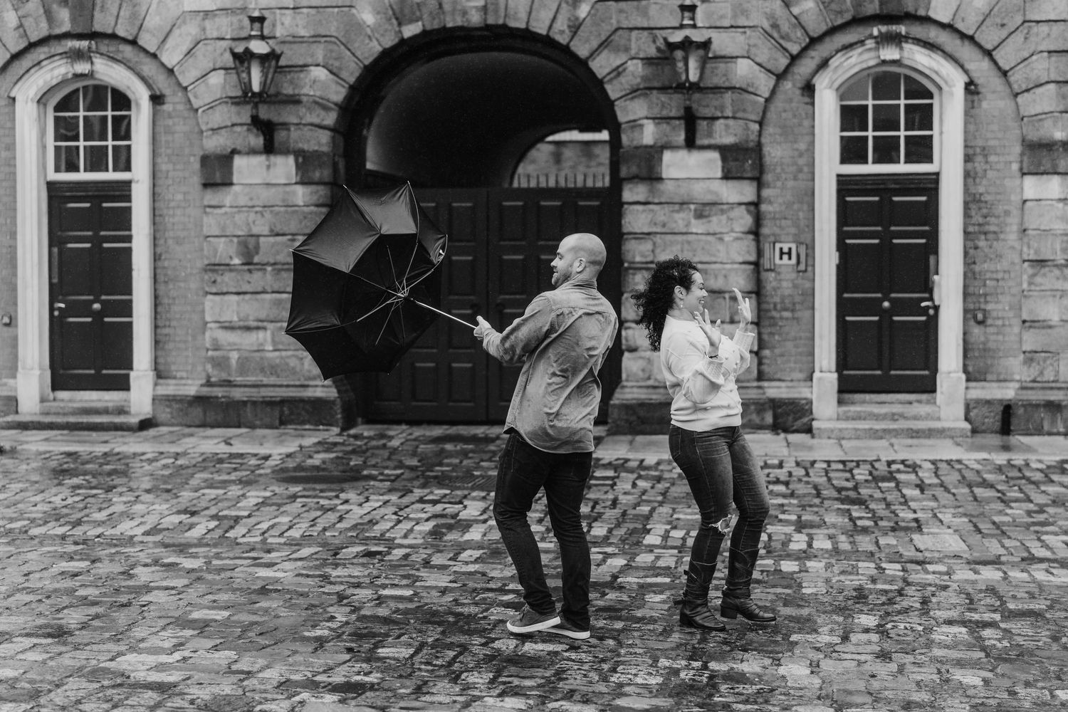 Black and white photo of a couple sharing an umbrella outside historic stone buildings. Proposal Dublin Photographer