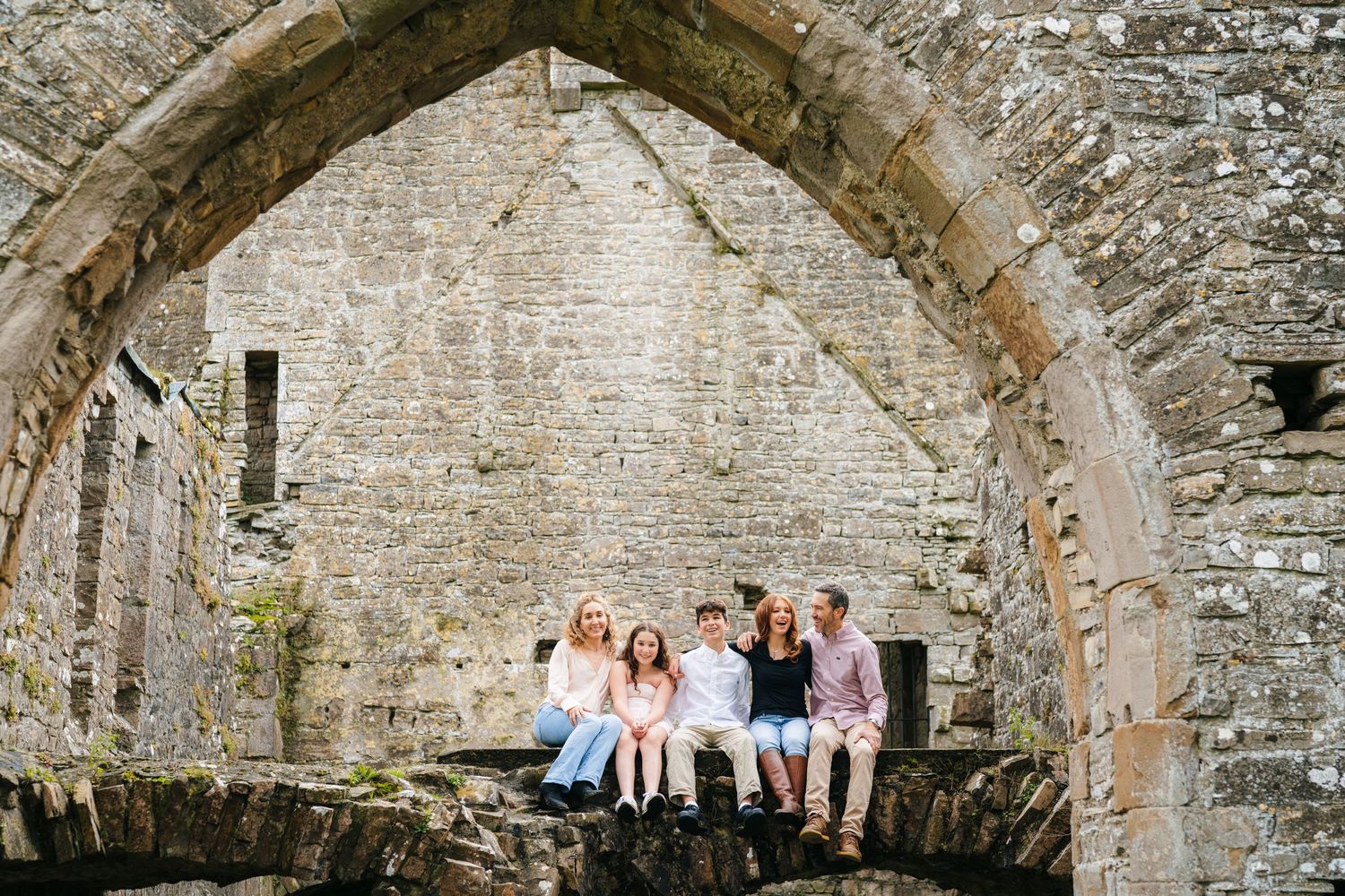 A group of people pose in front of a rustic stone wall with arches at an ancient castle or fortress. BEctive abbey family photographer dubli