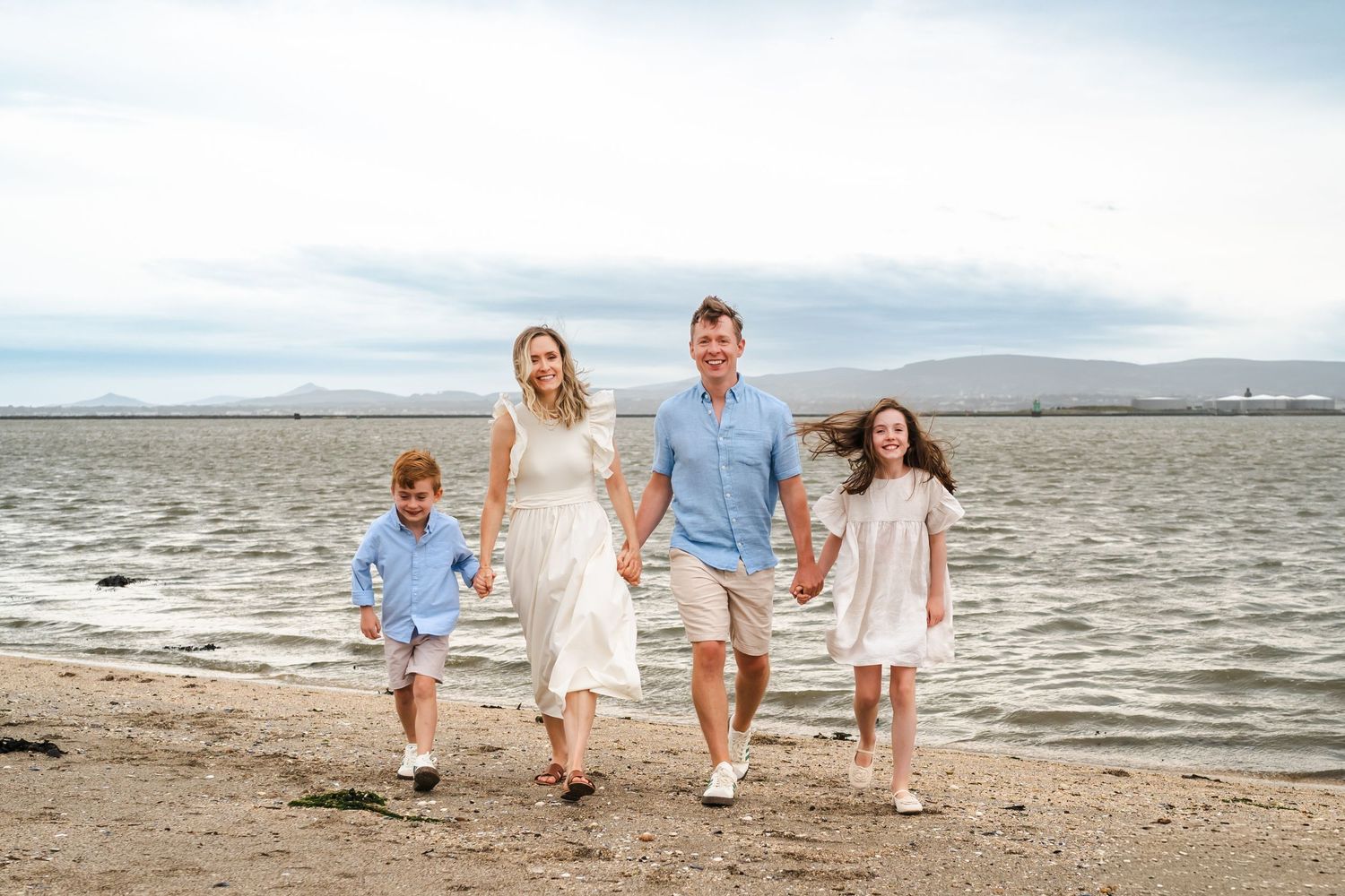 A family wearing white clothing walks together along a sandy beach shoreline during sunset.  Family photographer Dublin Ireland