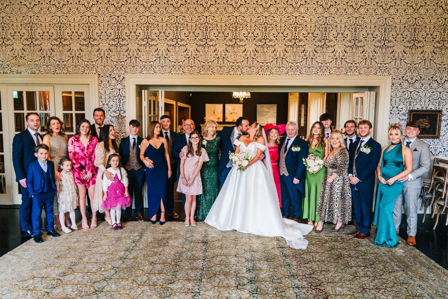Panoramic group photo of wedding guests and wedding party lined up in formal attire against patterned wall.  Virginia Park Lodge Wedding Pho