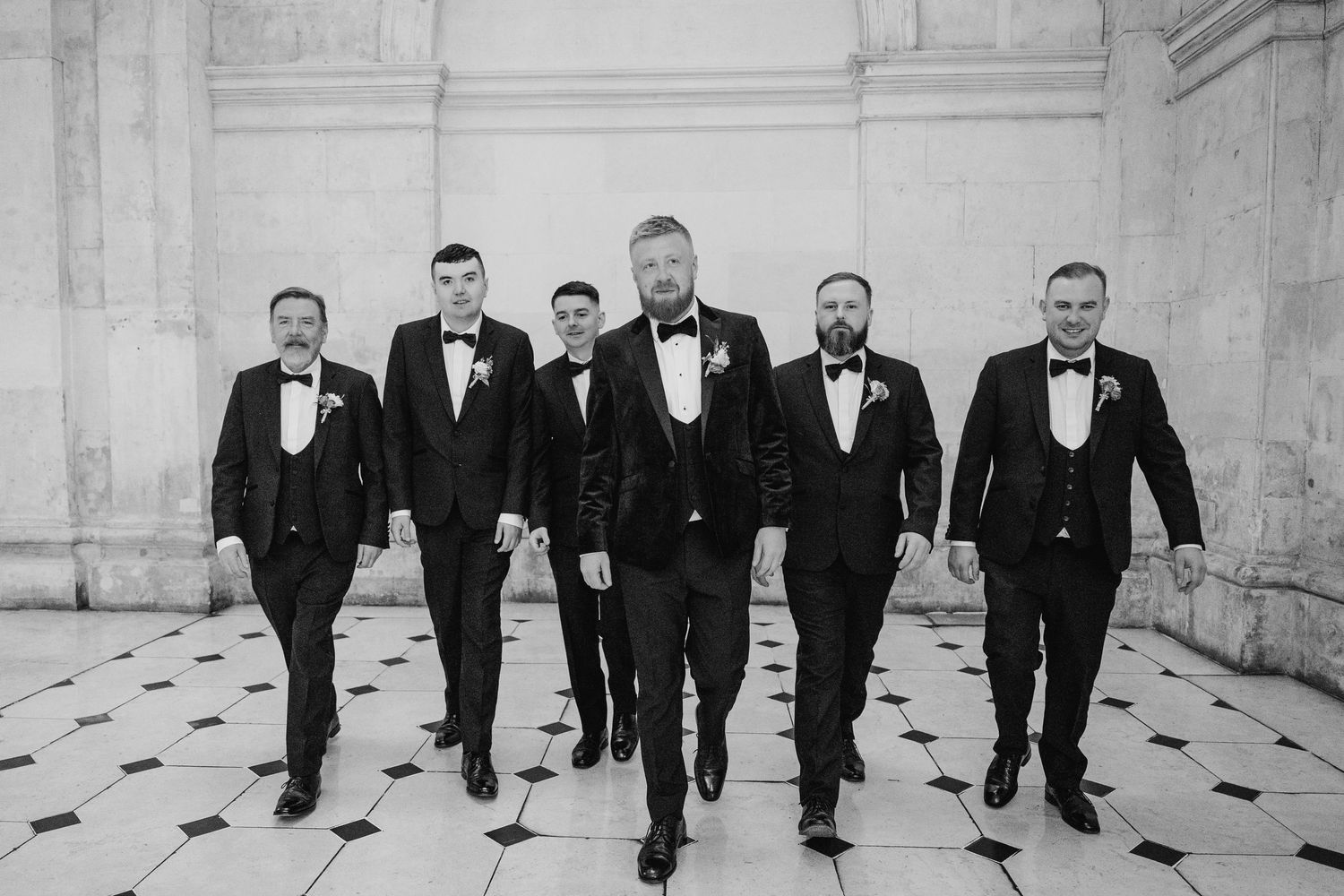 Groomsmen in black tuxedos lined up against a white stone building for formal wedding photos.Wedding photographer dublin