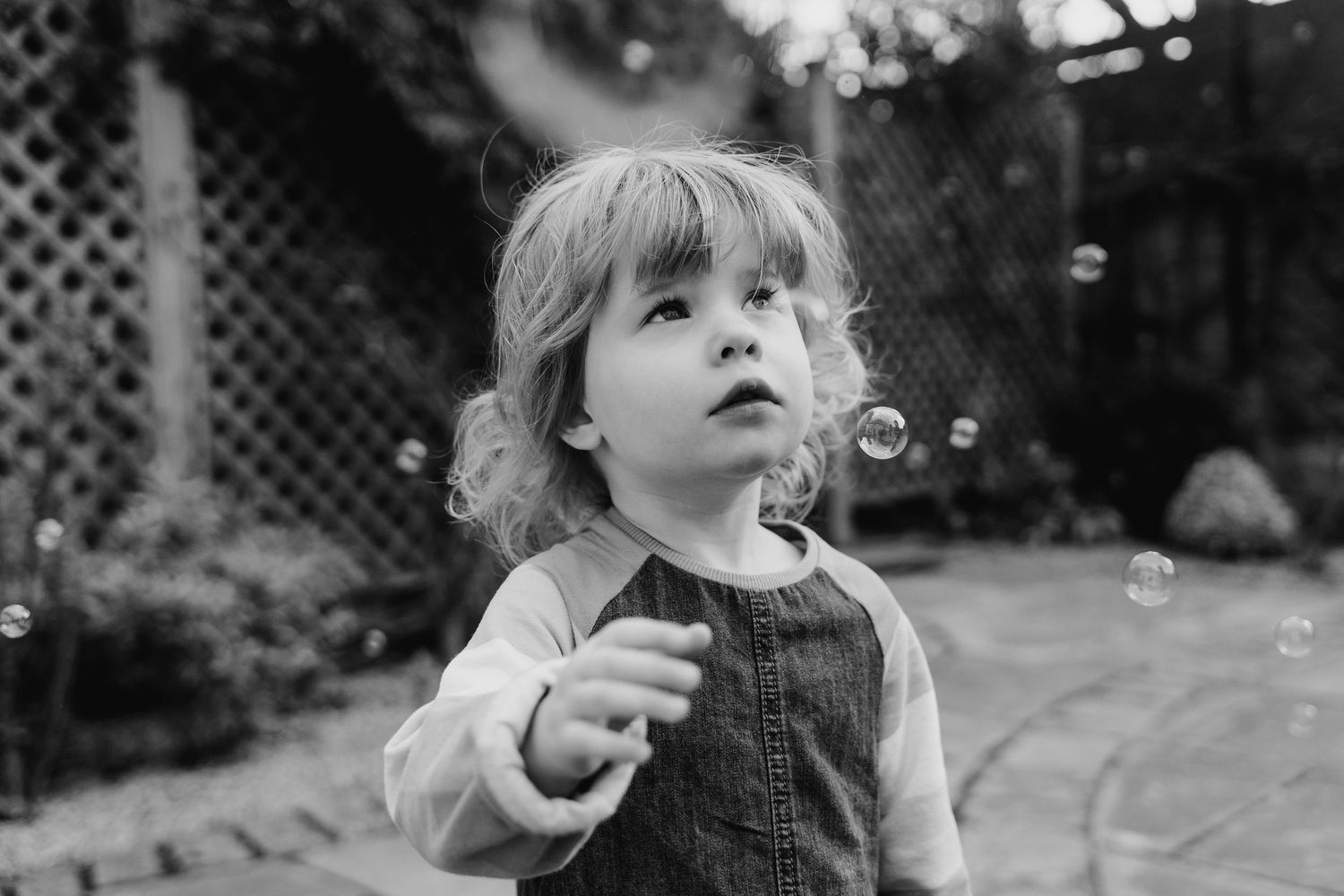 Child in black and white photography gazes up with wonder at floating soap bubbles in a backyard setting. family PHOTOGRAPHER Dublin