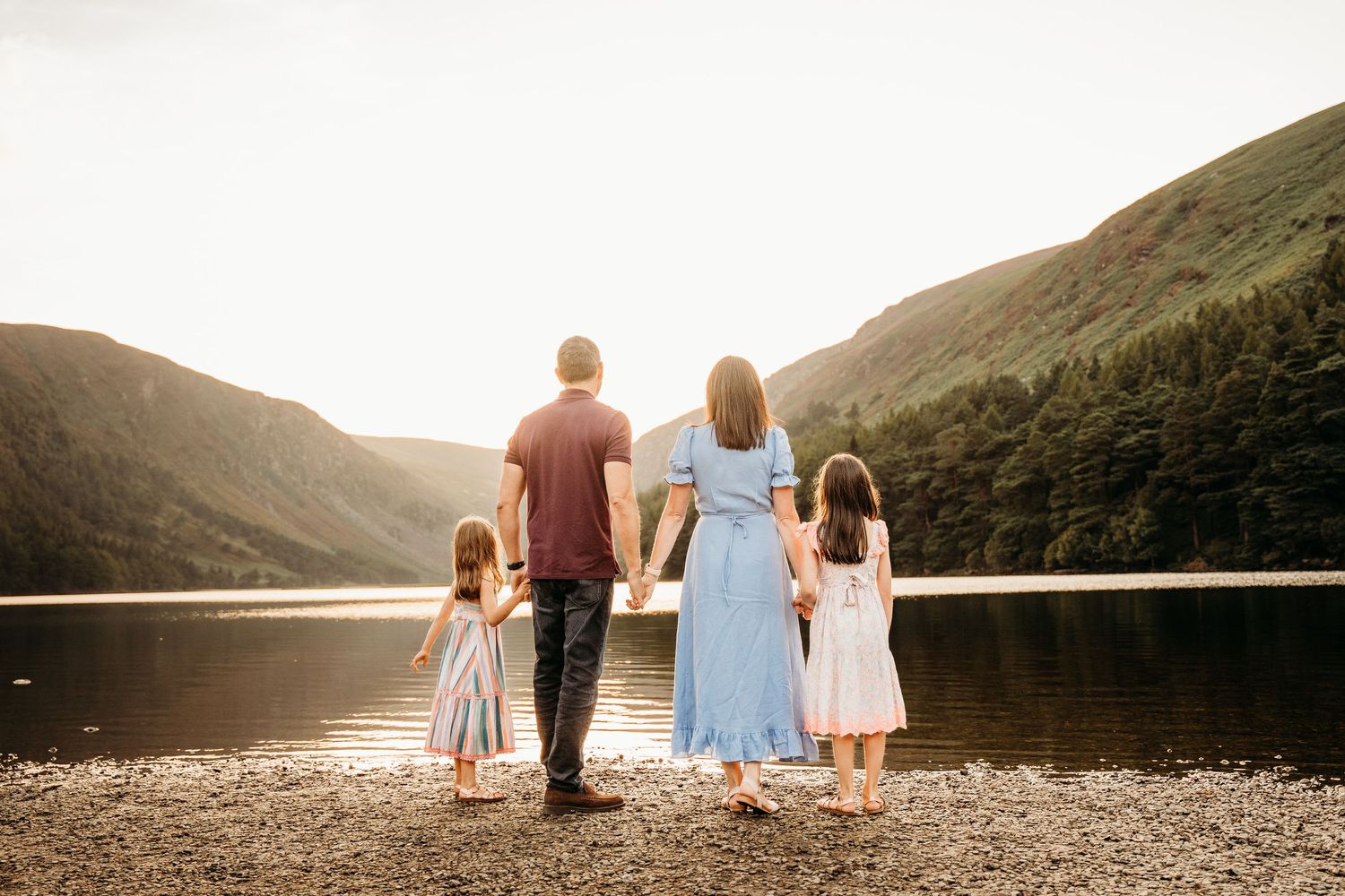 A family stands together holding hands at the edge of a serene lake surrounded by rolling hills at sunset. family PHOTOGRAPHER Dublin