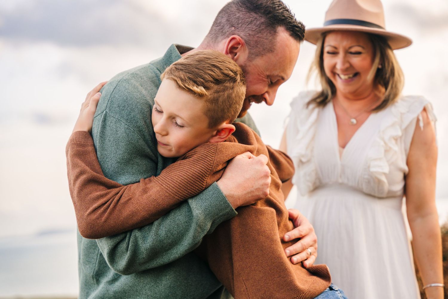 A tender family moment captured outdoors with loving embraces and a beige hat against a cloudy sky backdrop. family PHOTOGRAPHER Dublin