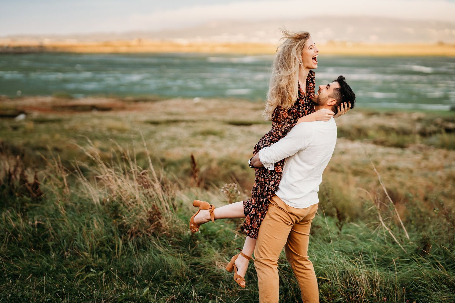 Couple embraces outdoors in windy grassland at sunset creating romantic silhouettes against golden sky. PHOTOGRAPHER Dublin ireland
