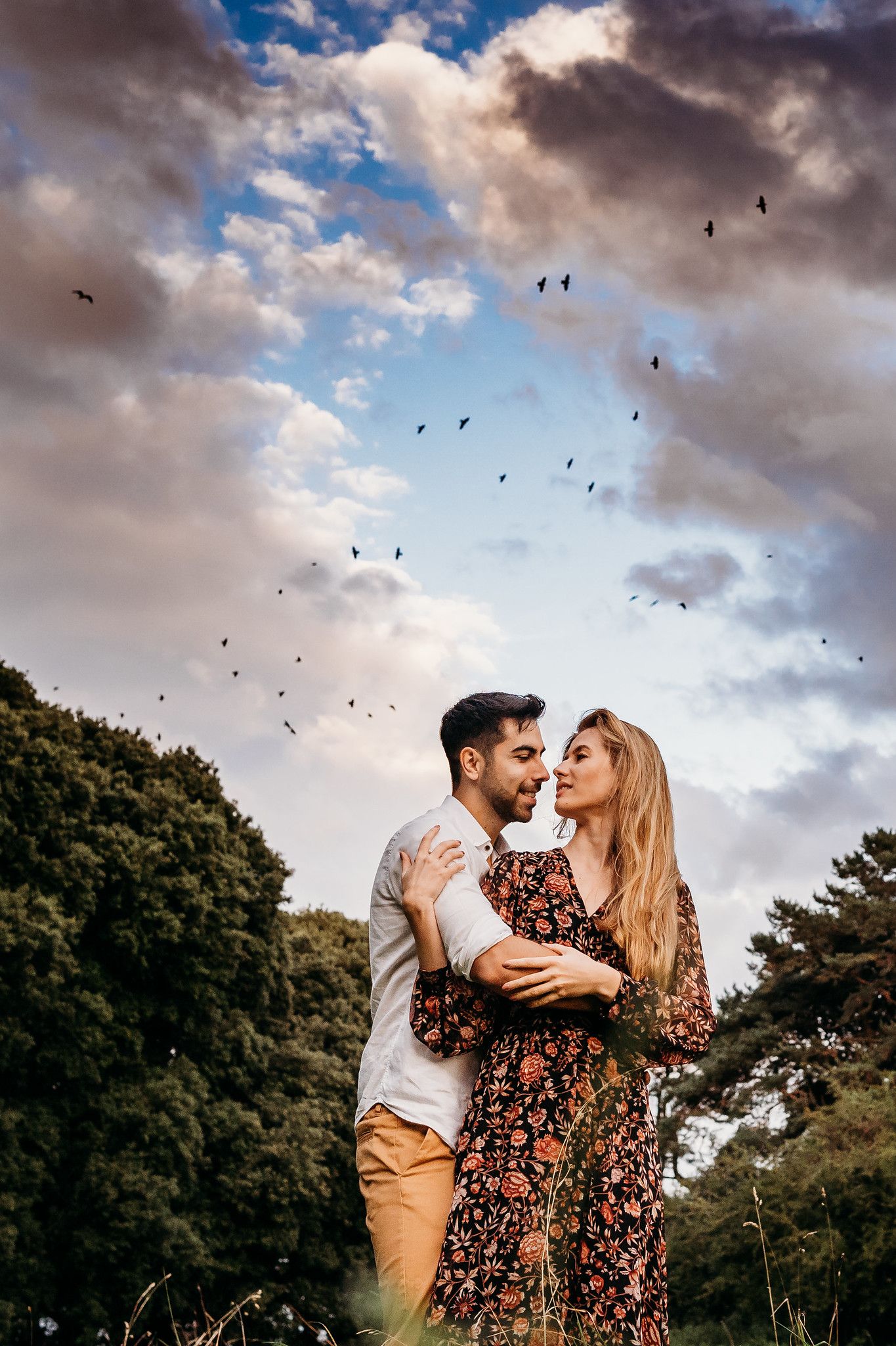 A couple embraces outdoors against a dramatic cloudy sky with birds flying overhead in a romantic photoshoot. PHOTOGRAPHER Dublin ireland