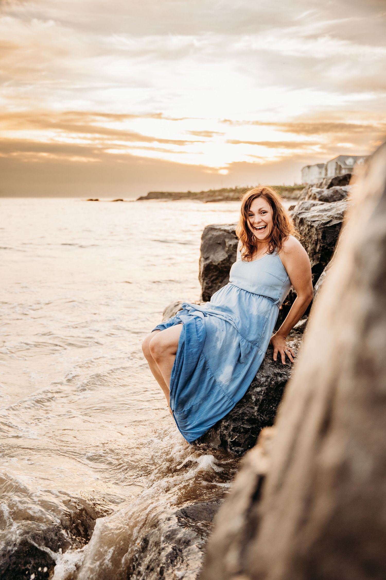 Person in flowing blue dress sits on coastal rocks at sunset while waves crash against rocky shore. PHOTOGRAPHER Dublin