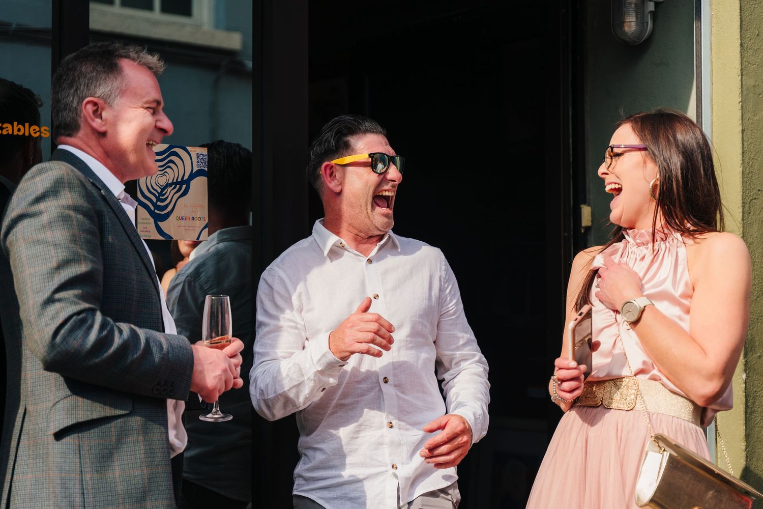 Three people share a laugh while holding drinks during a social gathering outdoors. Wedding PHOTOGRAPHER Dublin