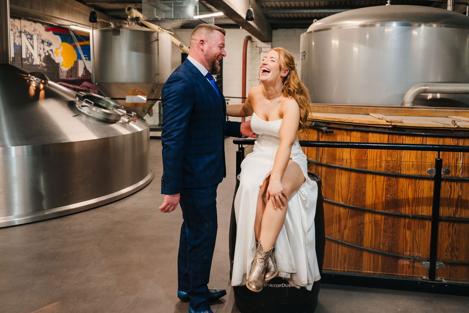 A newlywed couple shares a playful moment in a brewery setting with metal tanks and wooden barrels. Dublin wedding photogrAPher