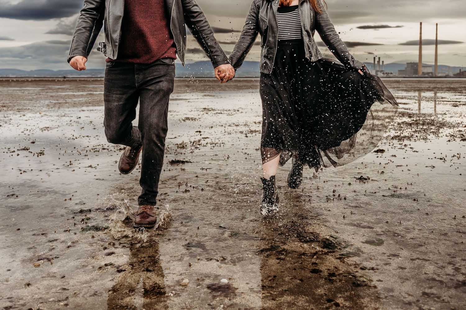 Couple splashing and playing in shallow tide waters under cloudy skies. engagement PHOTOGRAPHER Dublin