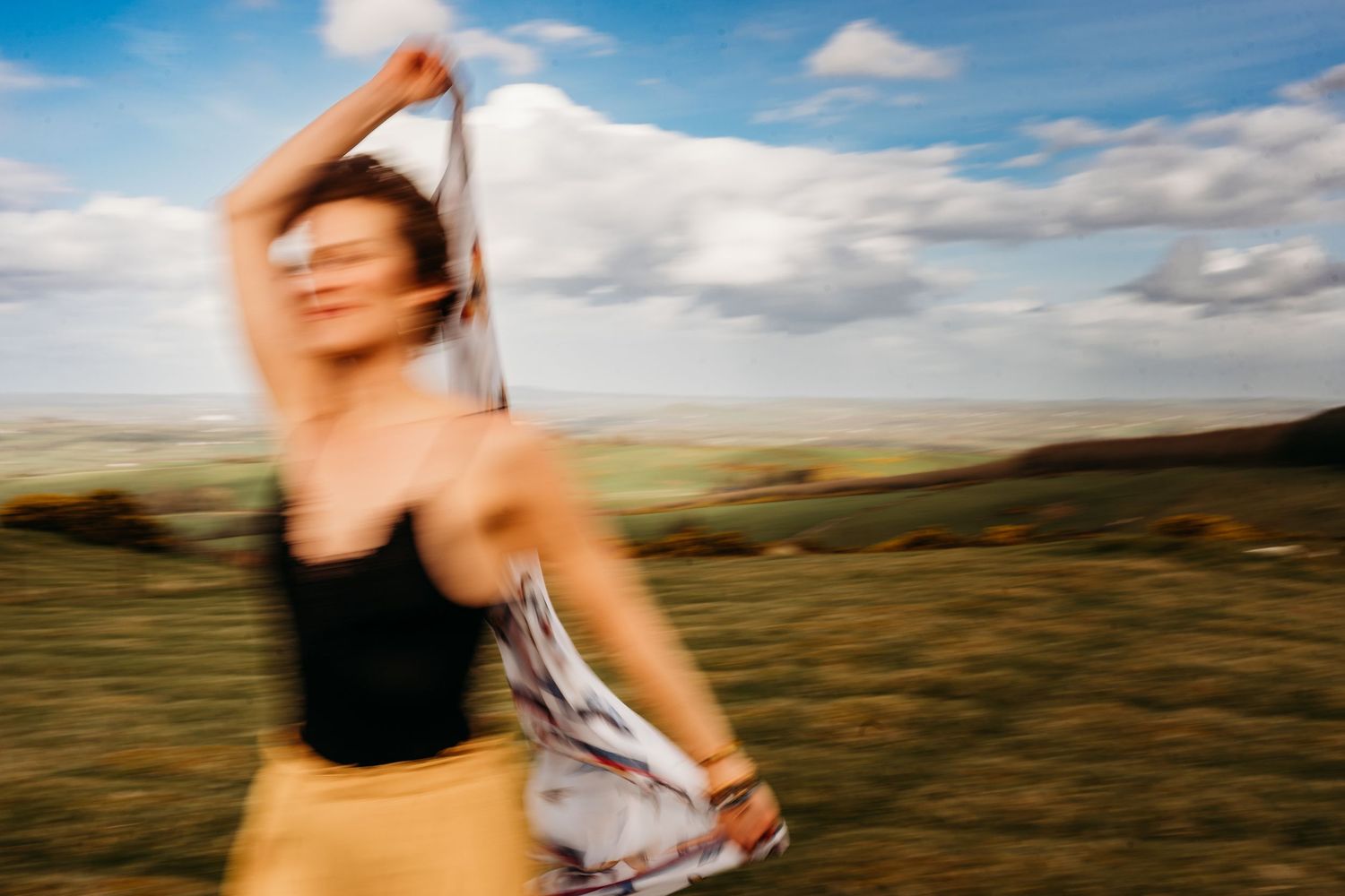 Motion blurred figure in black tank top dancing in a grassy field under blue cloudy sky. Portrait PHOTOGRAPHER Dublin