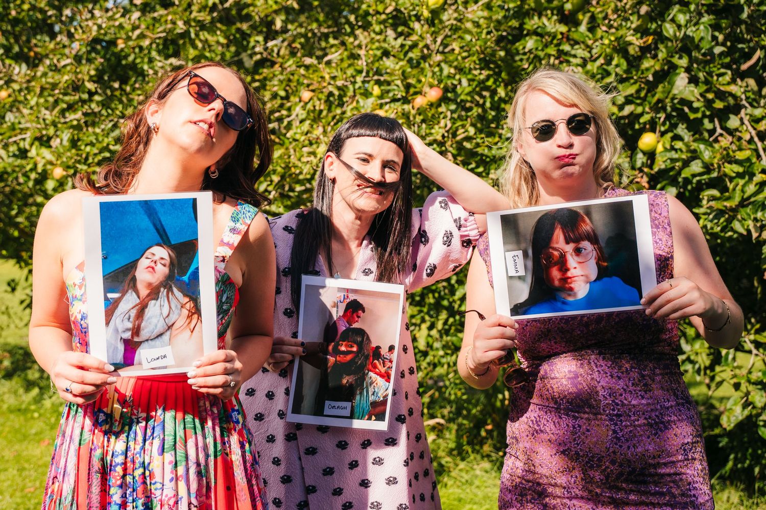 Three guests in colorful dresses pose outdoors while holding up old photographs. . Village Barn Tyrrellspass wedding photographer Ireland