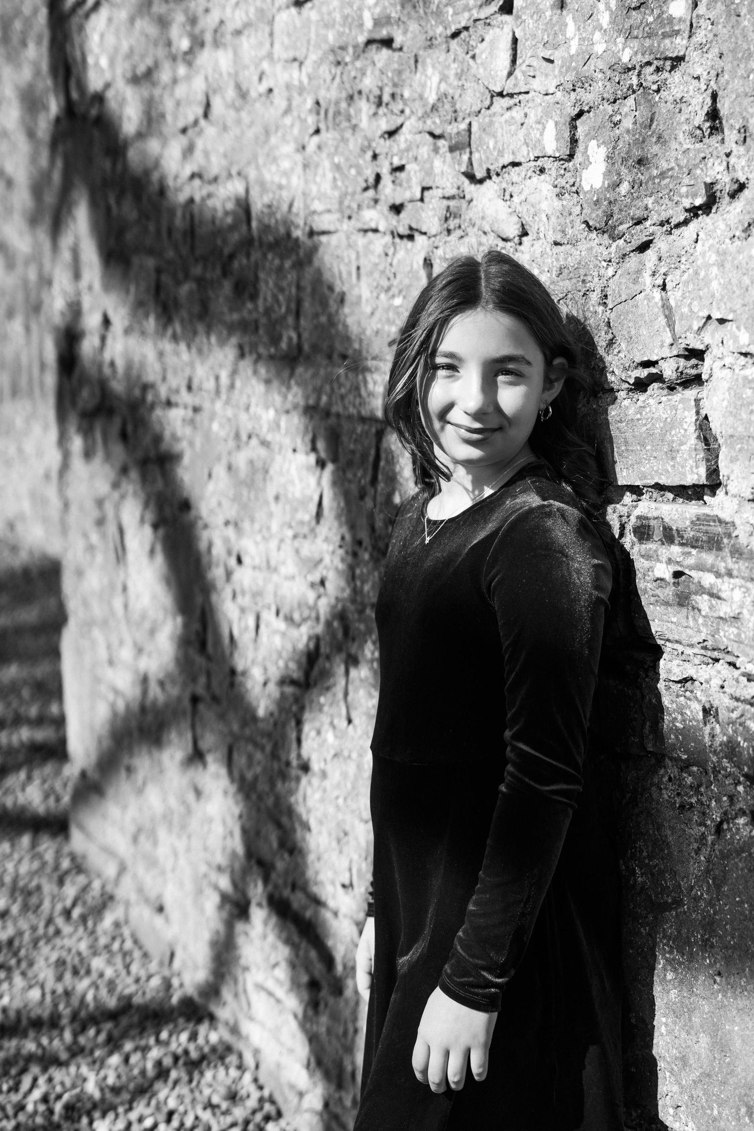 Black and white portrait featuring a velvet dress against an old stone wall with shadows. Family PHOTOGRAPHER Dublin