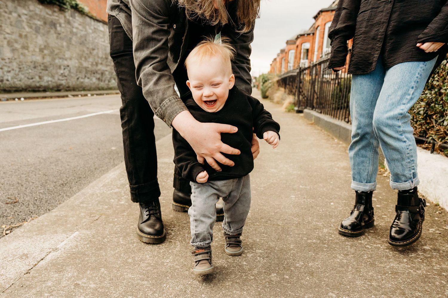 A toddler taking first steps on a sidewalk supported by adults on either side.  Family PHOTOGRAPHER Dublin