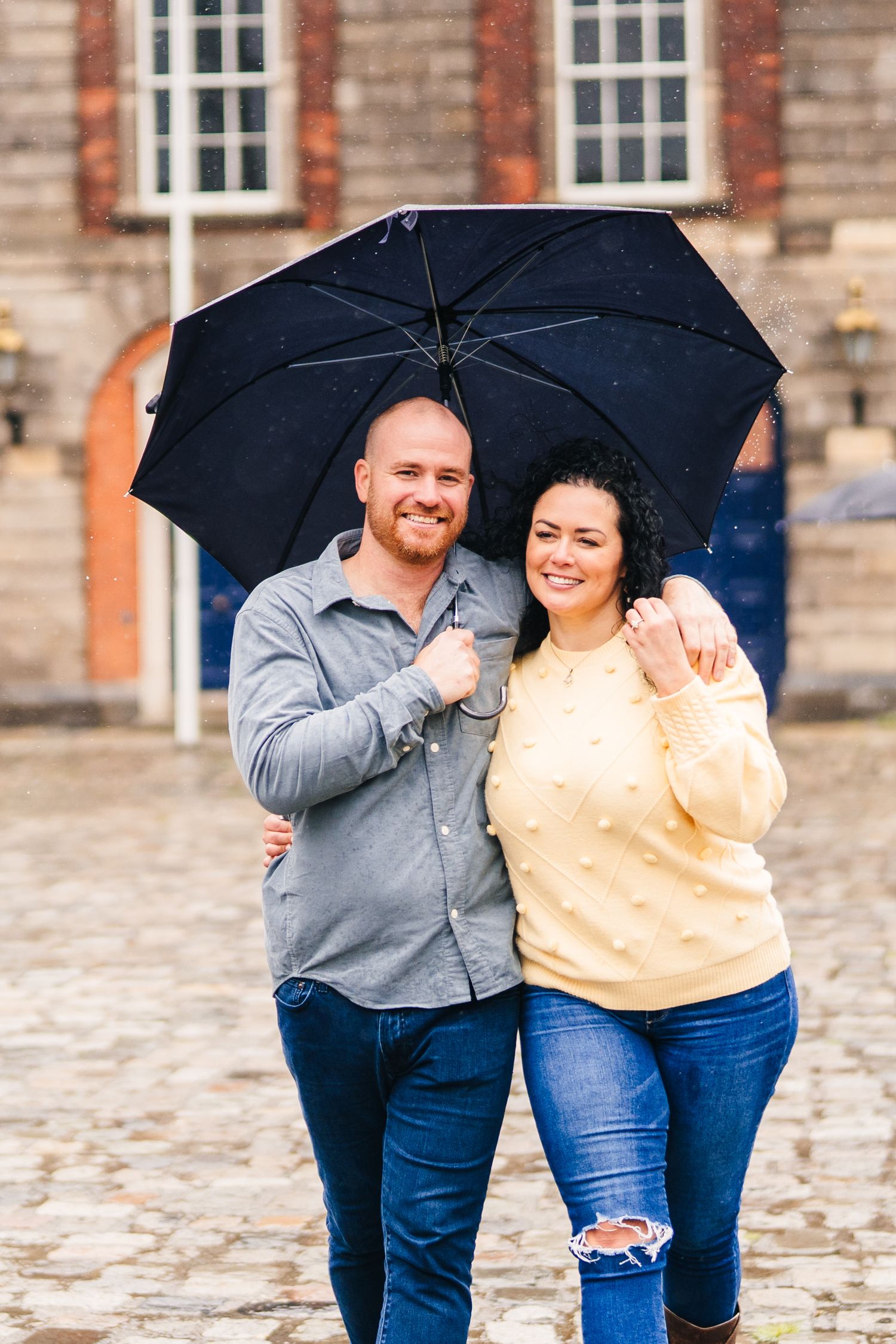 Couple sharing romantic moments under a black umbrella on rainy cobblestone streets. Proposal Dublin Photographer