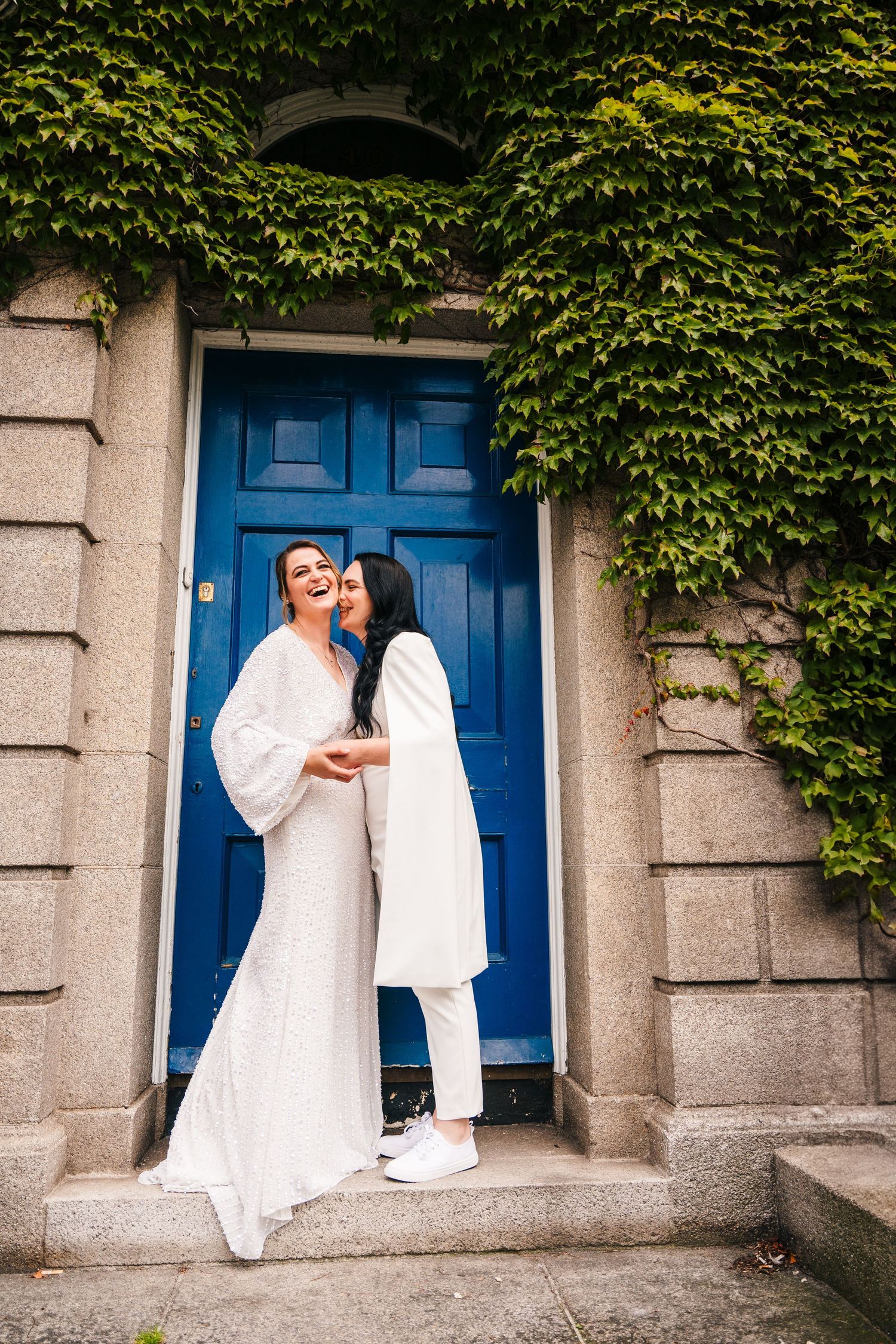 A couple in white outfits embrace in front of a blue door surrounded by ivy-covered stone walls. House Dublin Wedding Photographer